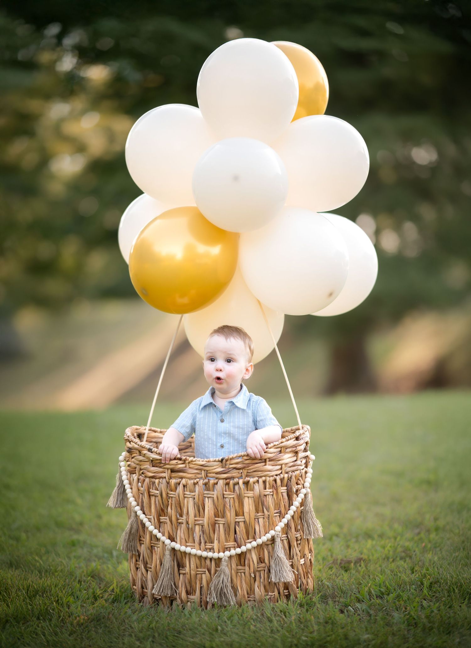 a little boy in a blue shirt stands in the jute basket of a toddler-sized hot air balloon with gold and white balloons floating overhead