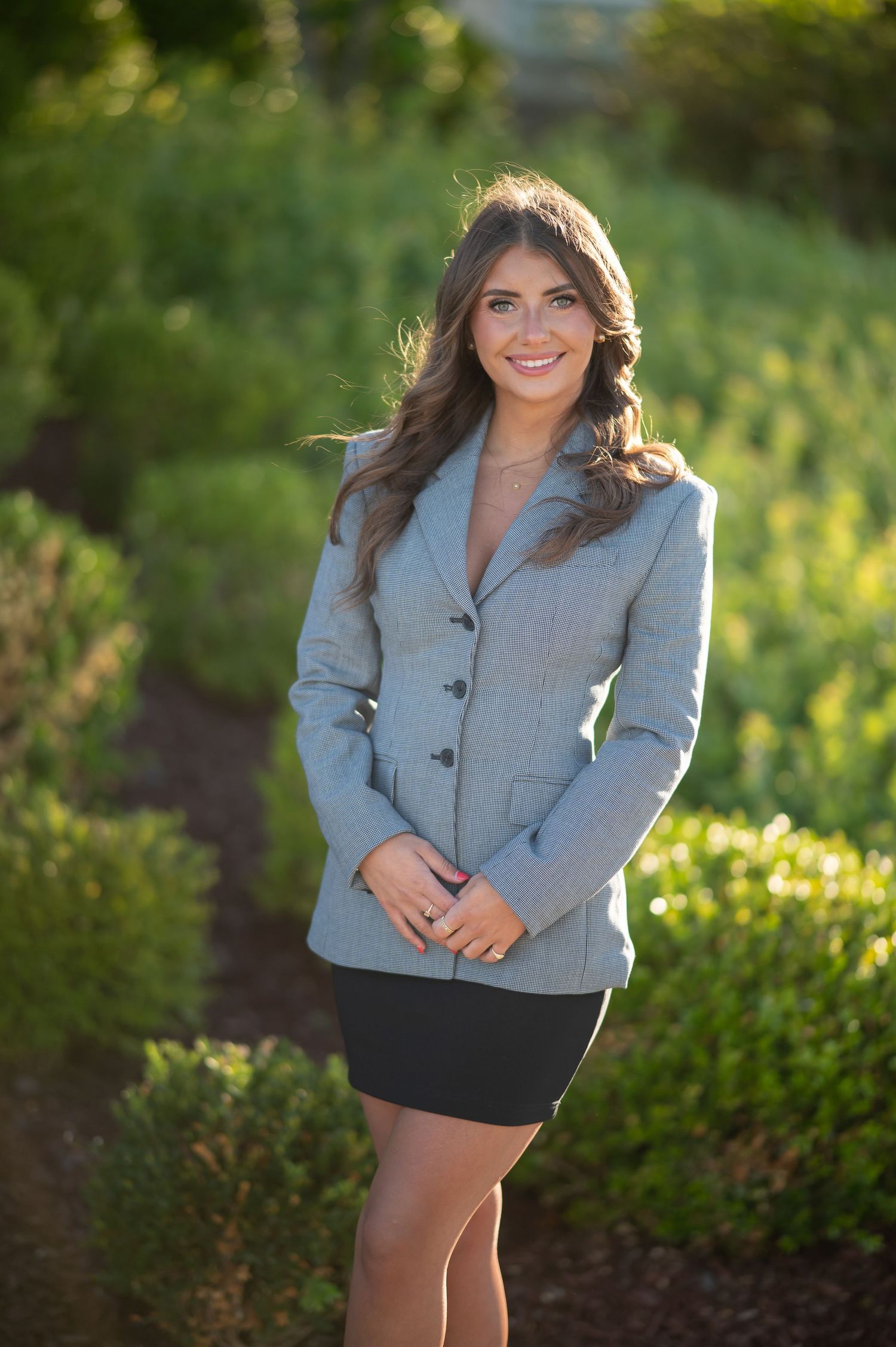 a young graduate student in a skirt and blazer poses and smiles for a senior portrait with green leaves in the background