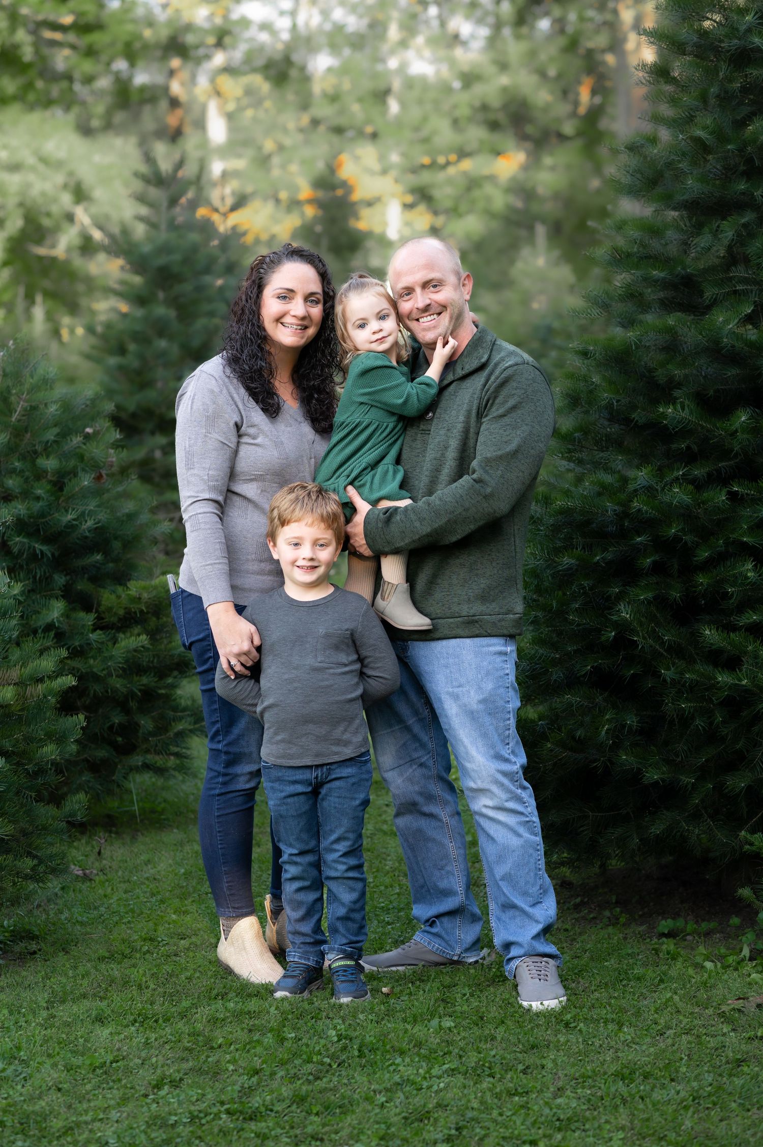 a family of four dressed in muted greens and grays poses in front of a row of Christmas trees at a tree farm in Fairmont, WV