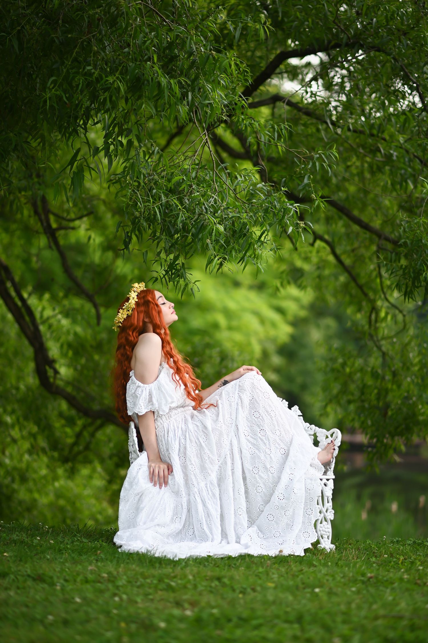 a woman in a long white dress with vibrant red hair sits on a white iron bench surrounded by lush trees and grass