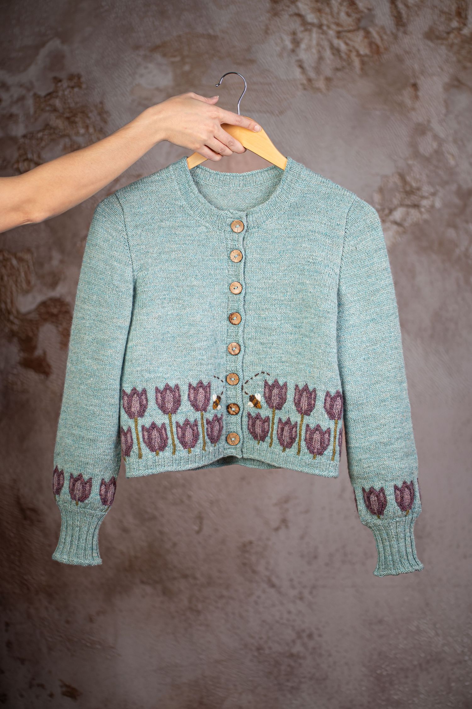 Fashion photography – a woman's bare arm extends into the frame holding a wooden hanger with a hand knit teal sweater on it