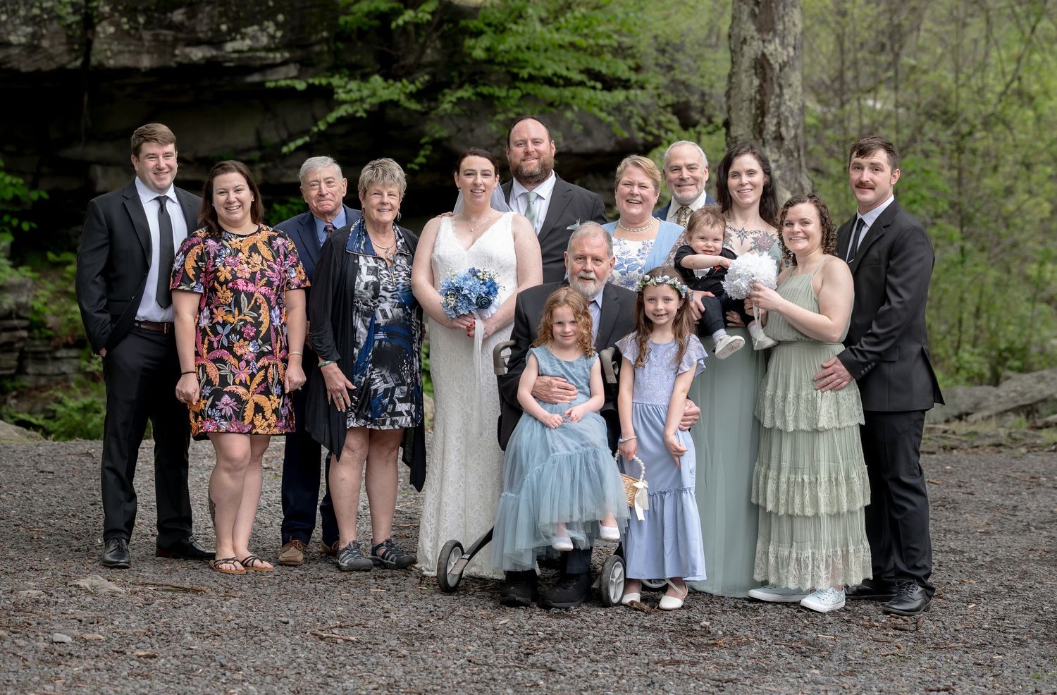 an extended family in wedding attire pose around a happy bride and groom at an outdoor wedding, with rocks and trees in the background