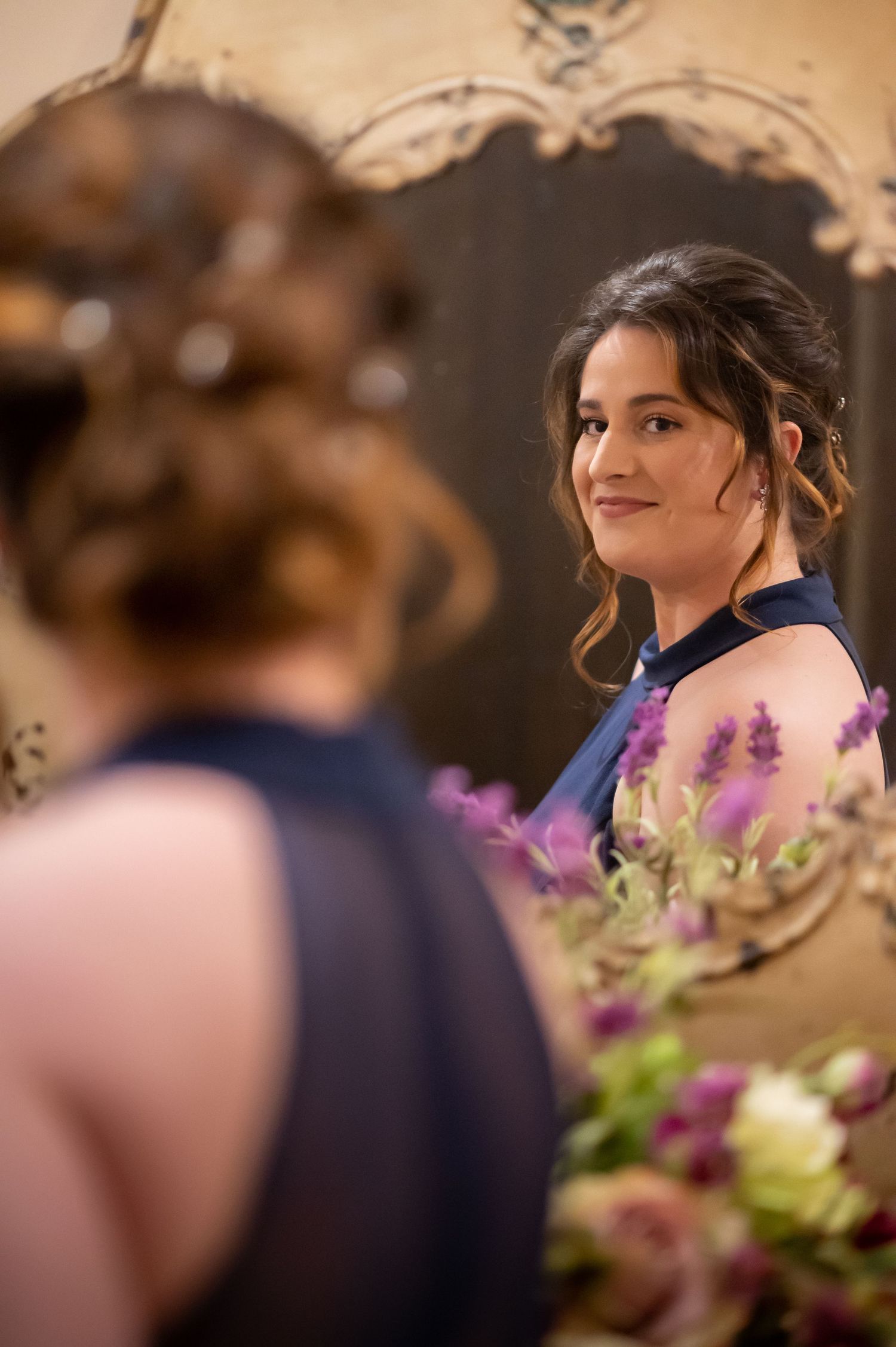 a bridesmaid in a navy blue dress smiles at her reflection in an ornate golden mirror as she finishes getting ready for her friend's wedding