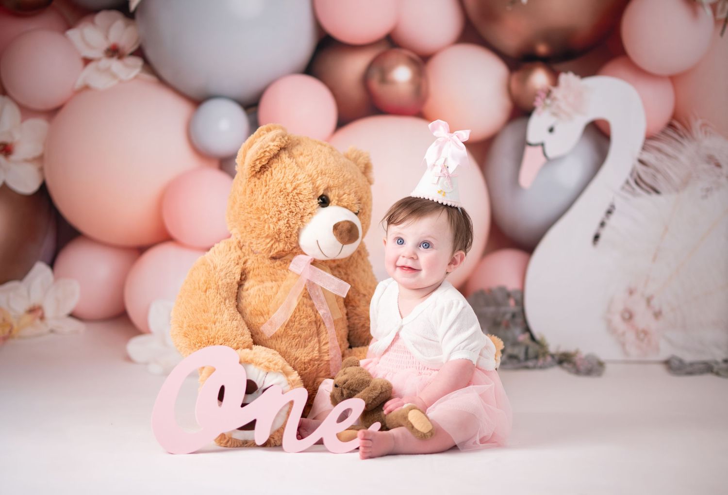 a little girl in a pink dress and birthday hat poses with her teddy bear for first birthday portraits with pastel balloons in the background