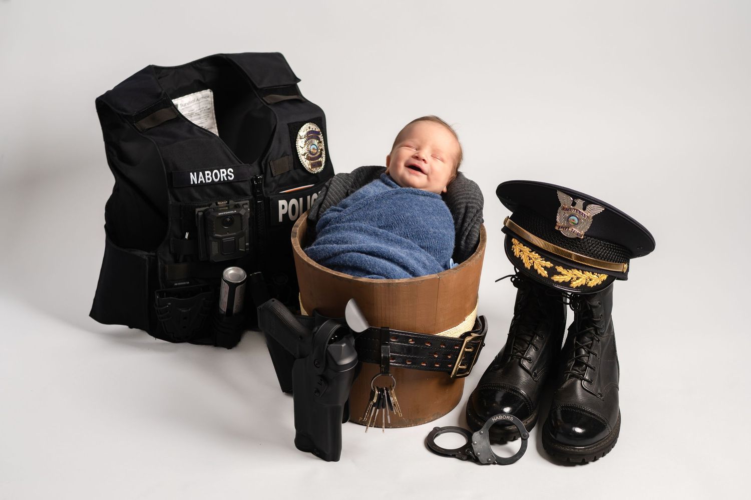 Newborn baby smiling in a wooden bowl surrounded by police gear including vest, hat, boots, and handcuffs.