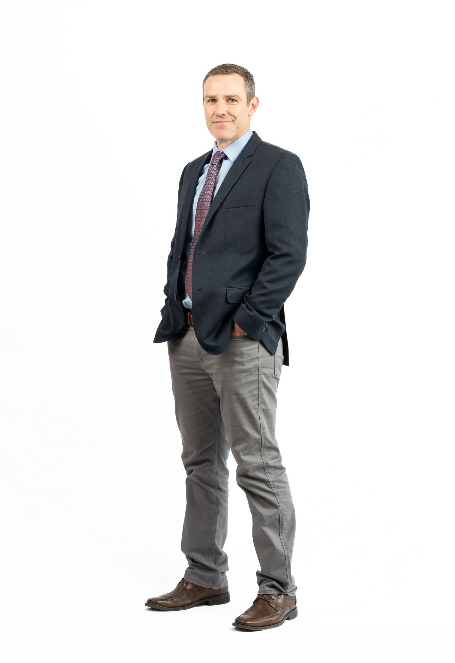 a man in a navy and gray suit poses with his hands in his pockets for a full-body headshot