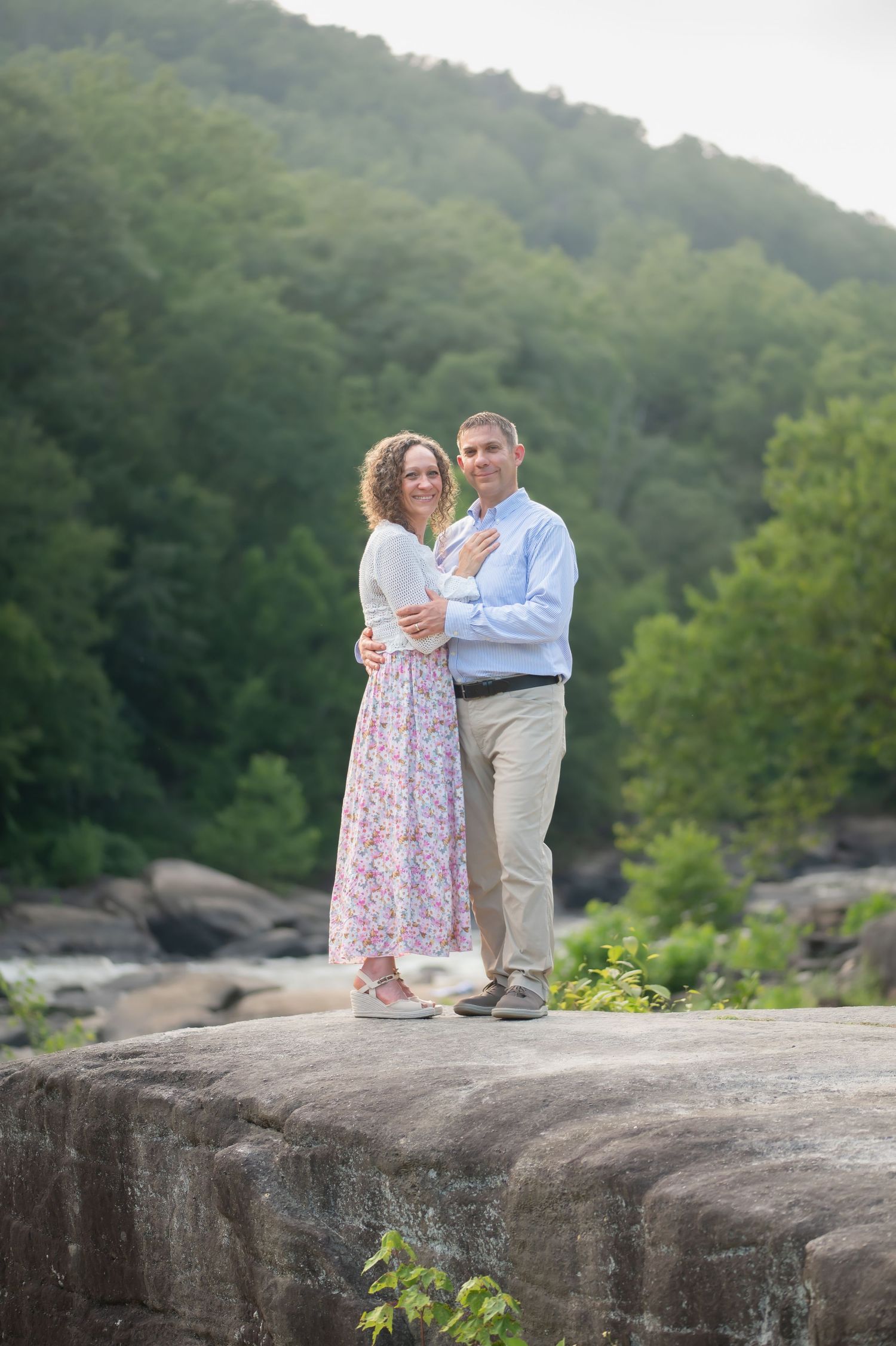 a husband and wife in muted pastel colors embrace and pose for a portrait on a large rock in front of rocks and waterfalls in Valley Falls