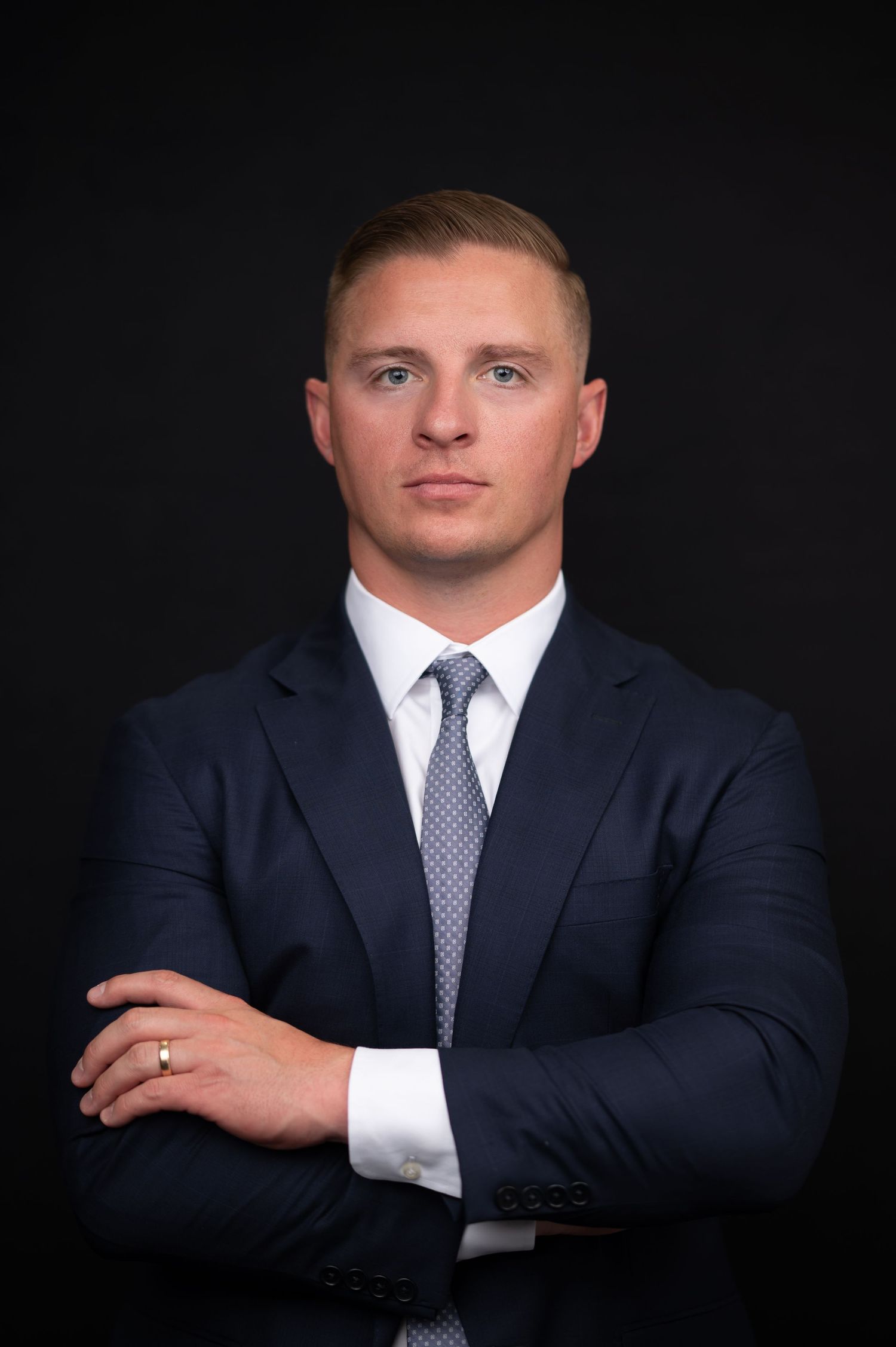 a dramatic professional headshot of a man in a dark navy suit jacket and light blue tie, looking into the camera with a serious expression