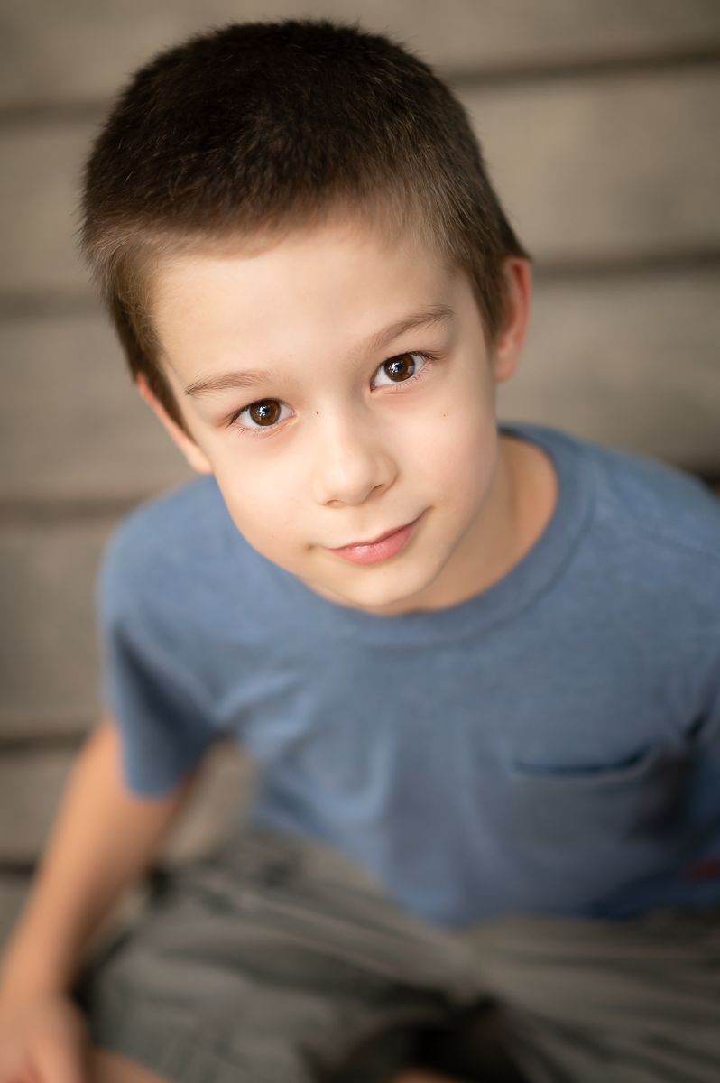 a little boy in a blue shirt poses sitting cross-legged and smiles up at the camera