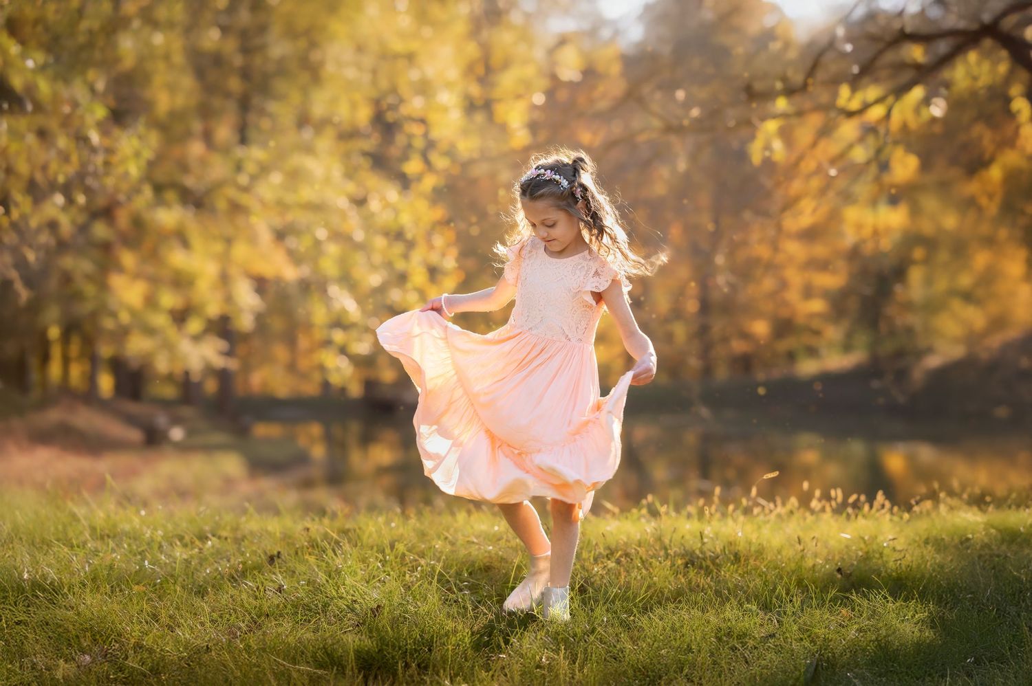 a little girl in a fluffy pink dress twirls in a golden sunlit field with autumn trees in the background during a portrait session