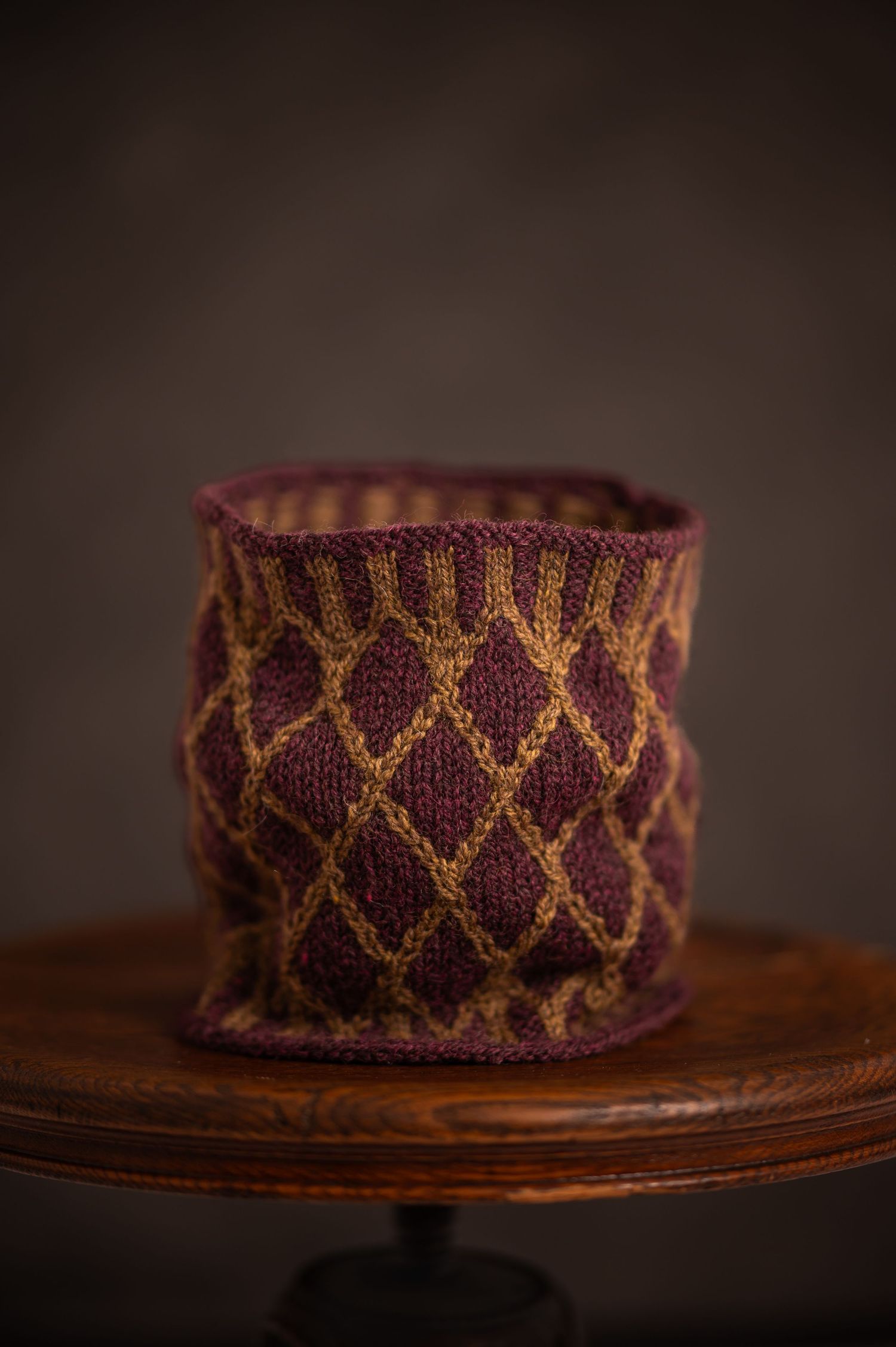 product image of a burgundy and gold hand-knitted cowl standing on an old wooden stool with a deep brown background