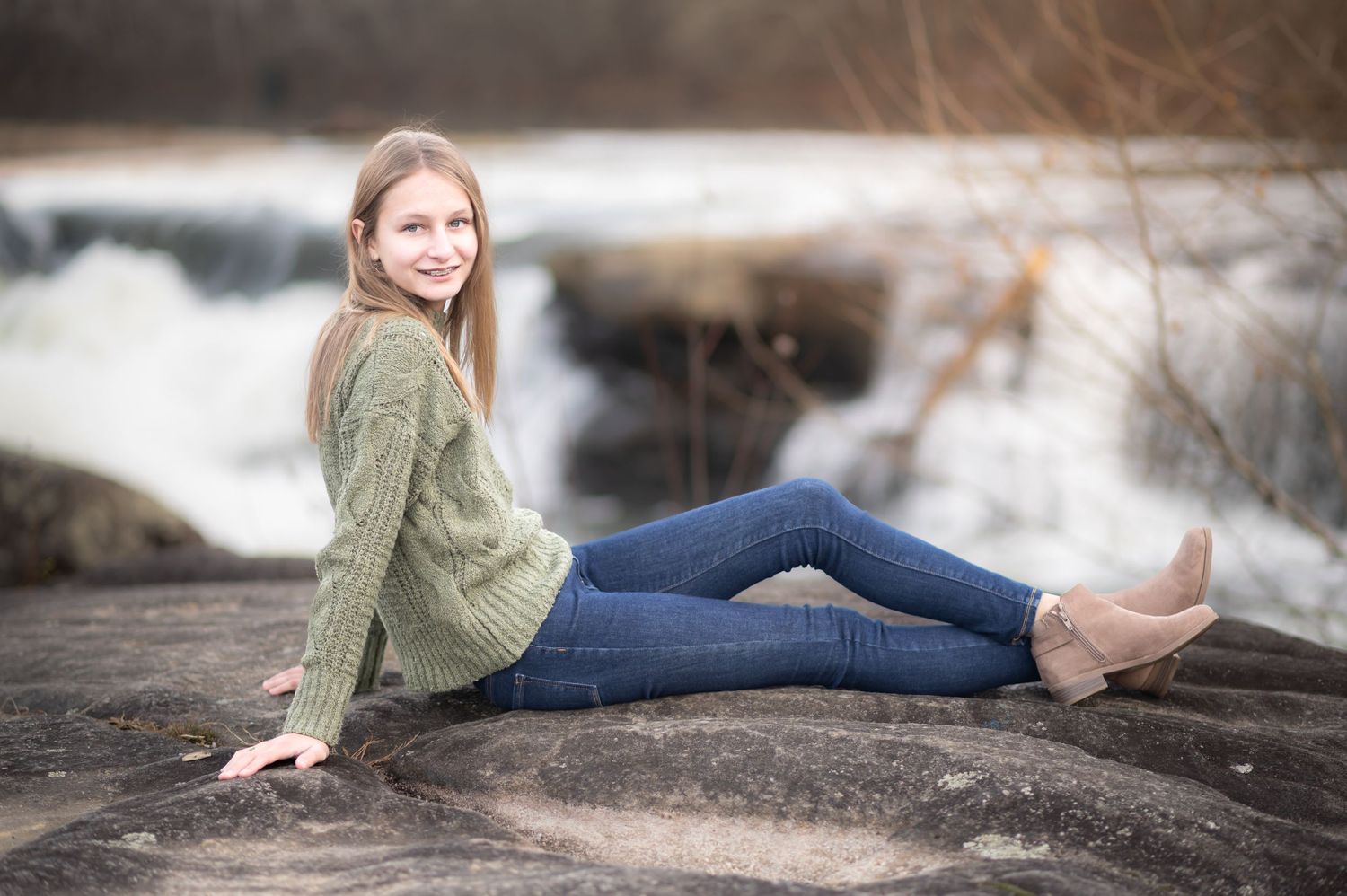 a young woman in a green sweater and jeans poses seated on a large rock, with roaring waterfalls in the background