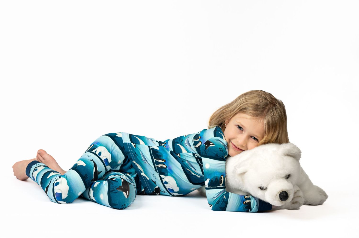 a high-key product photo of children's bamboo pajamas; a little blonde girl lies holding a stuffed bear on a bright white studio background