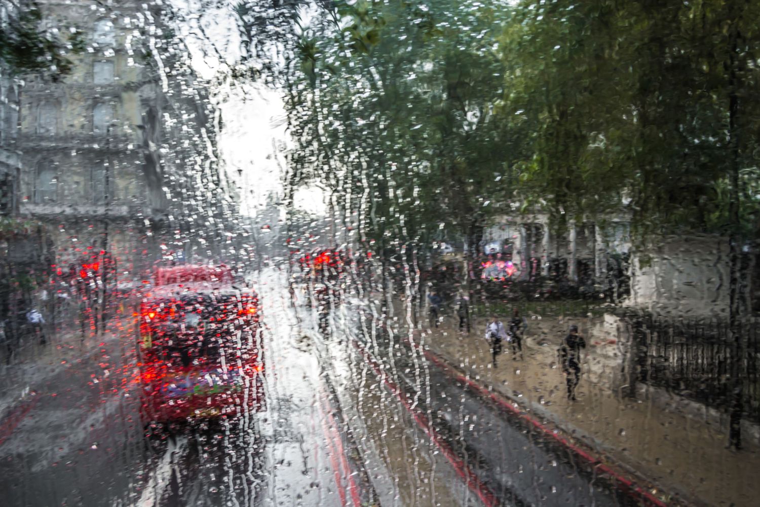 Rainy day view through a wet glass window of a London street with red buses and pedestrians walking.