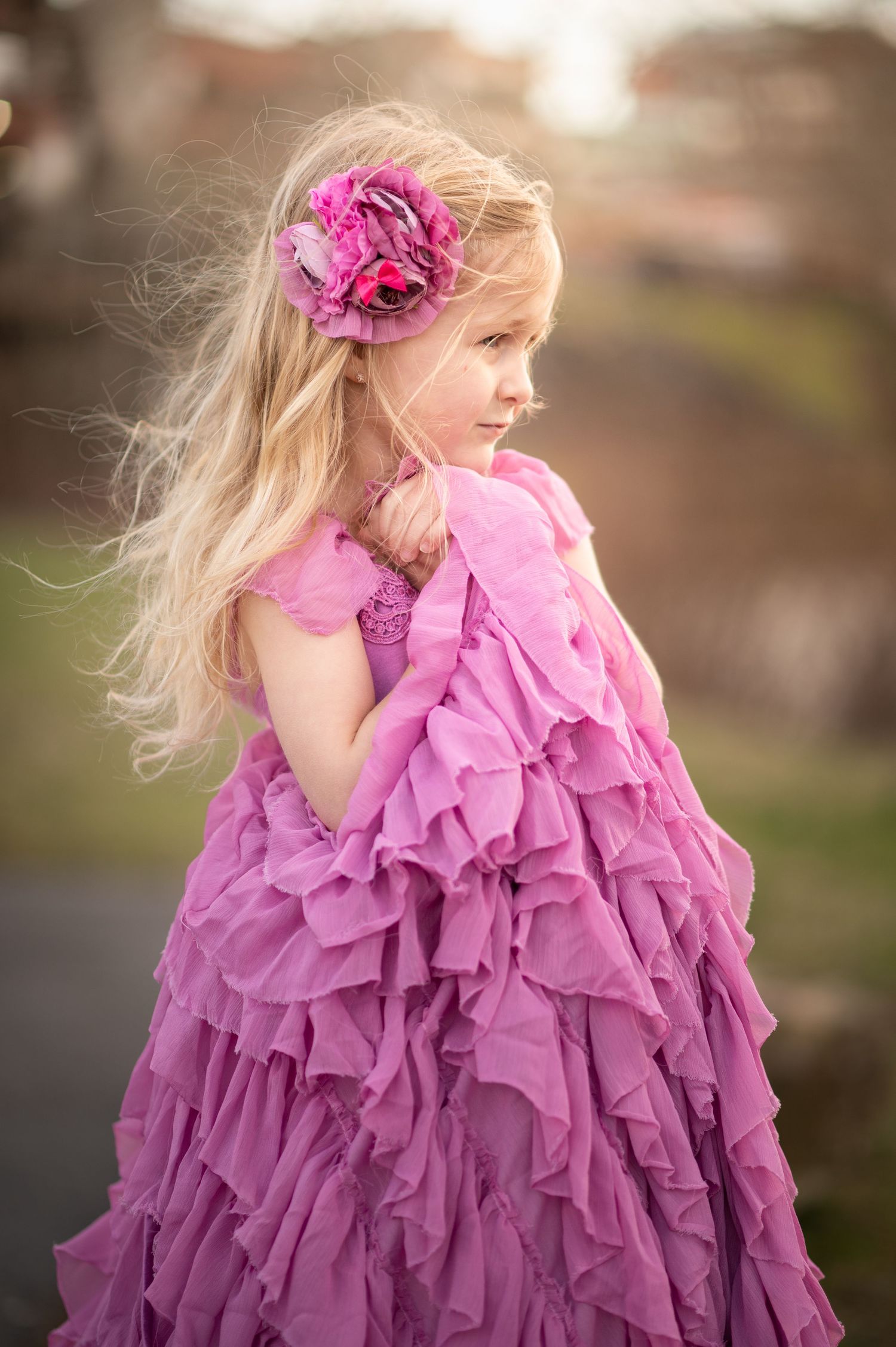 a little girl in a frilly mauve dress stands in front of a river as the breeze blows her blonde hair