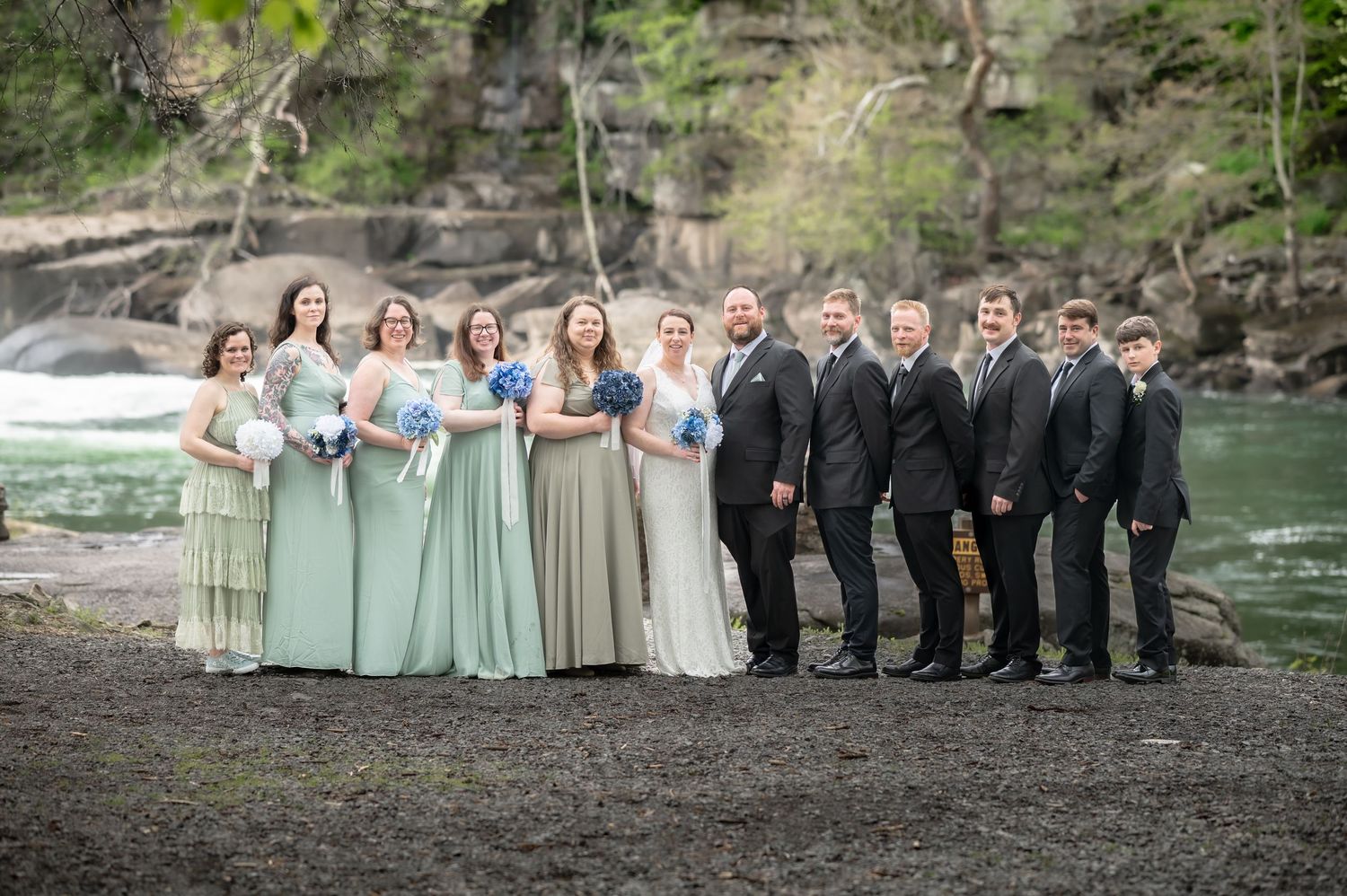 a full bridal party in mint green and black pose in front of the waterfalls at Valley Falls State Park for a wedding portrait