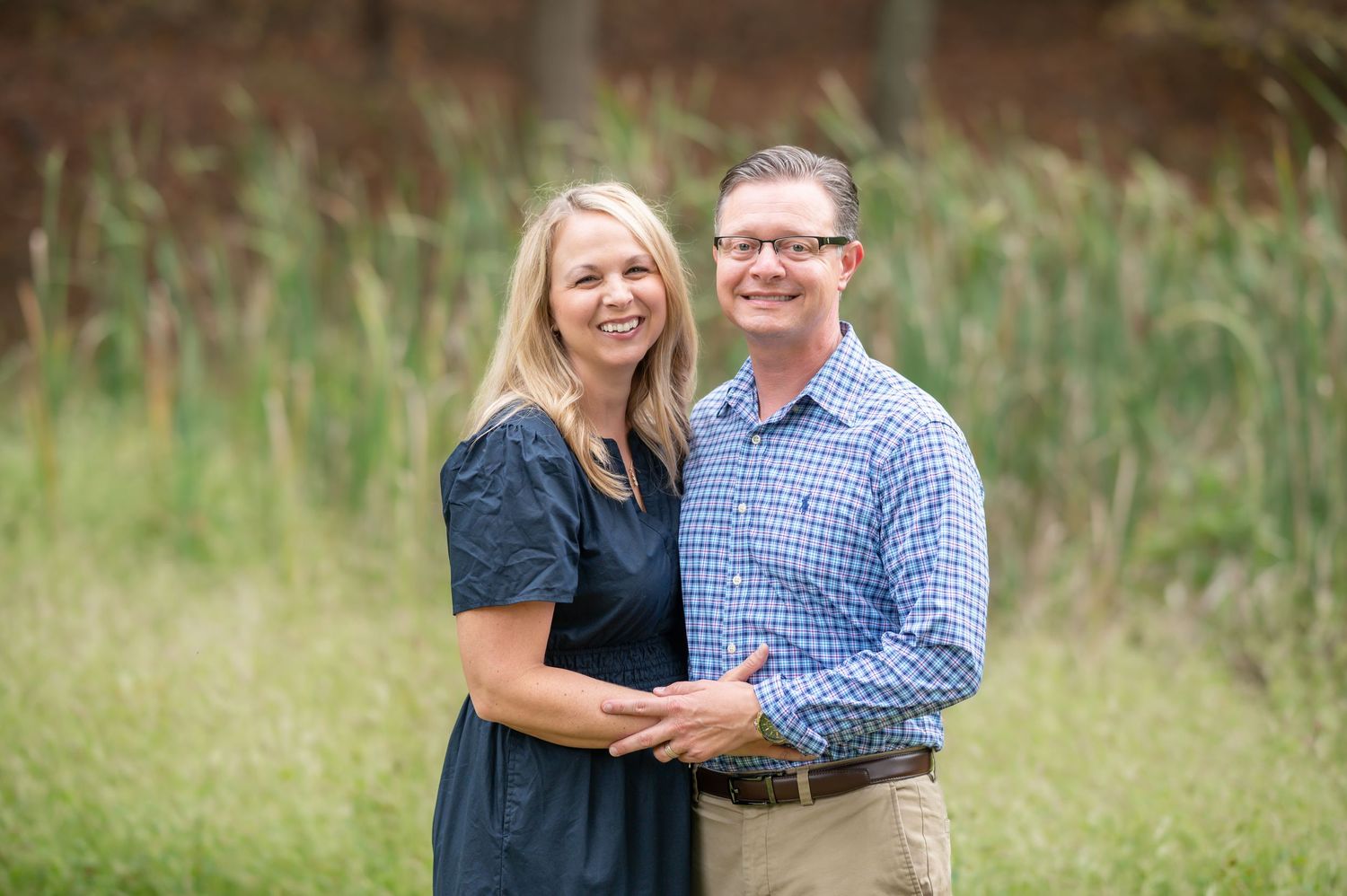 a husband and wife stand with arms around each other and smile for a portrait, with green sunlit grass blurred in the background