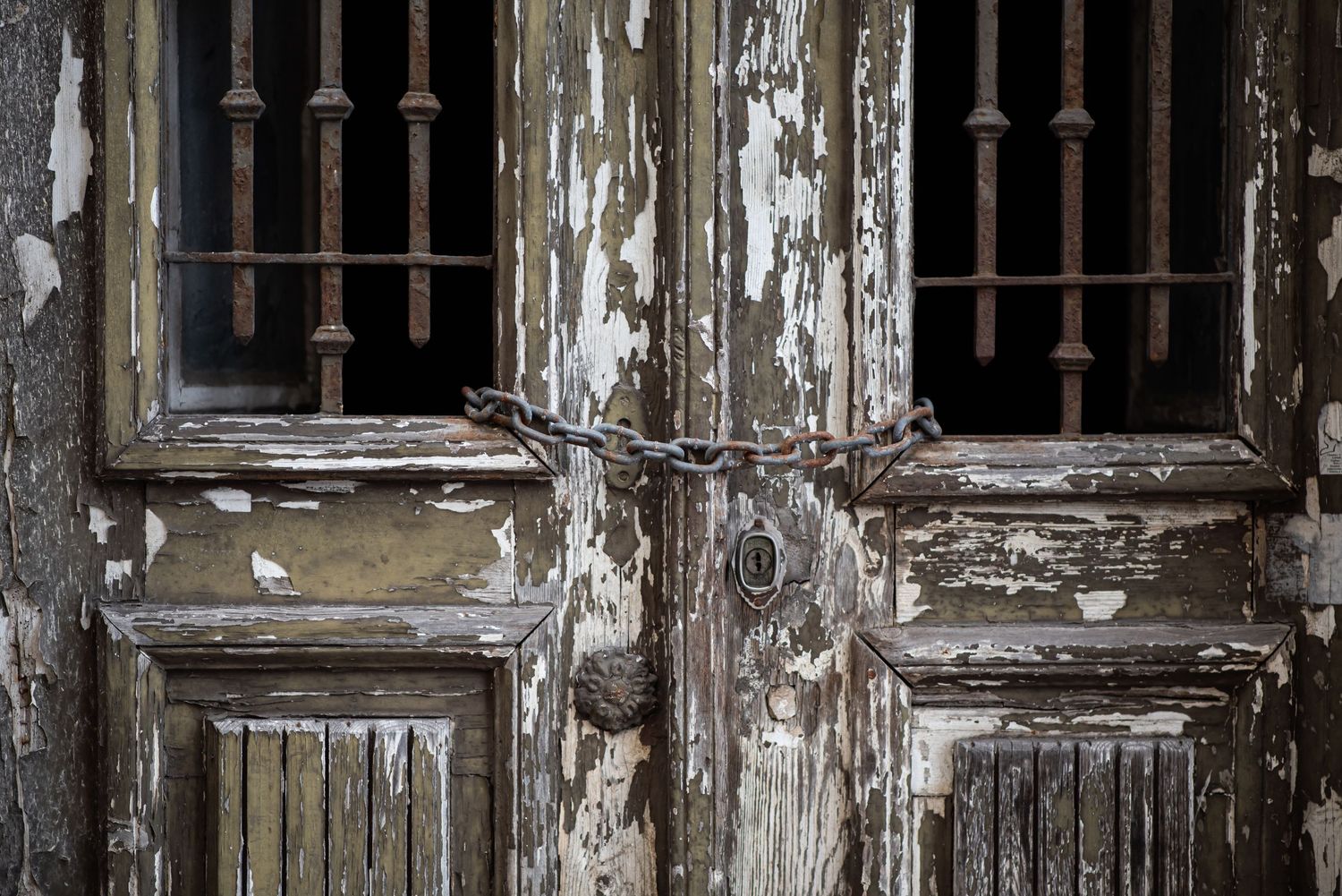 Old wooden double doors with peeling paint, rusty chain lock, and barred windows showing age and decay.