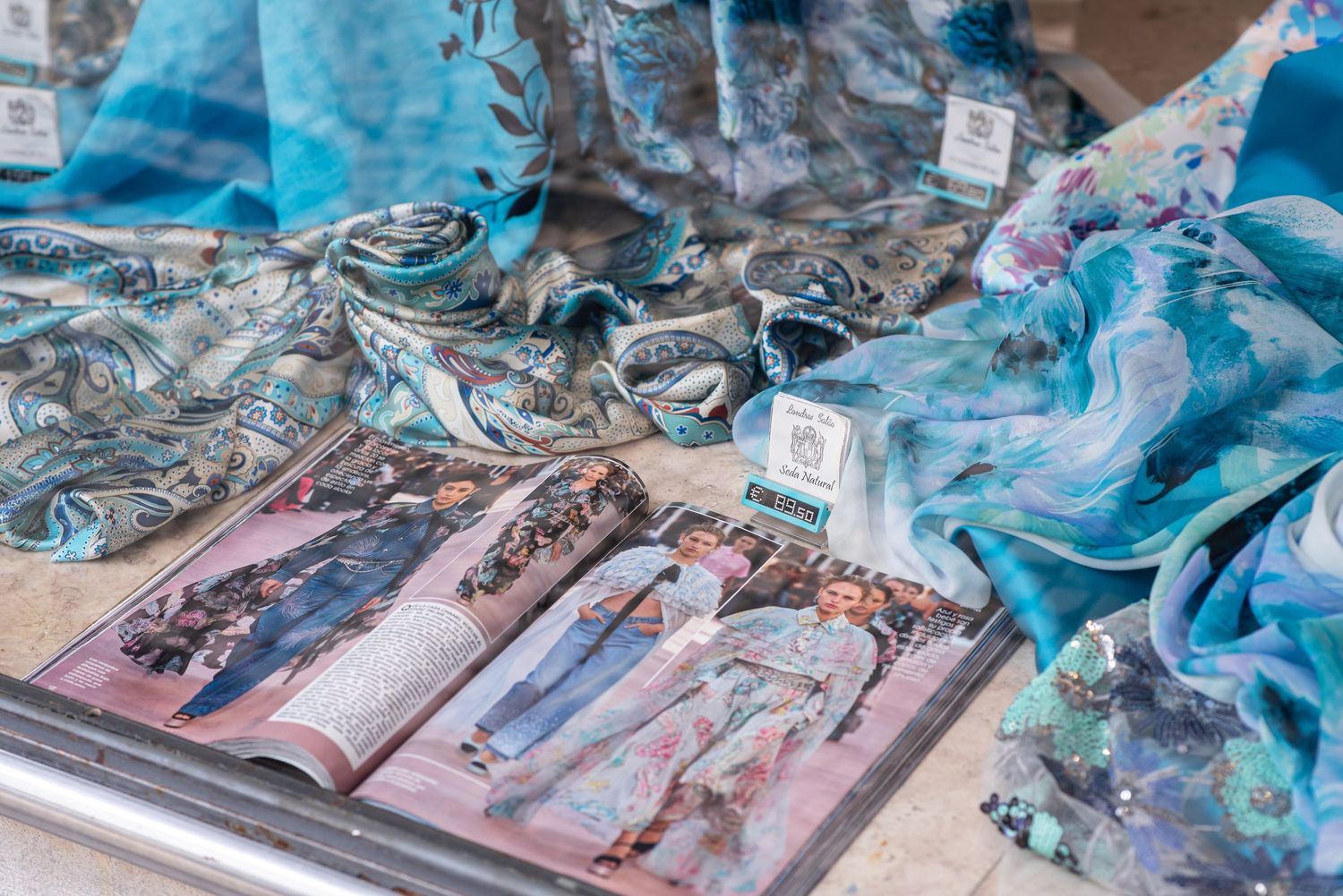Elegant blue silk scarves and fashion magazine displayed on a table at a boutique market stall.
