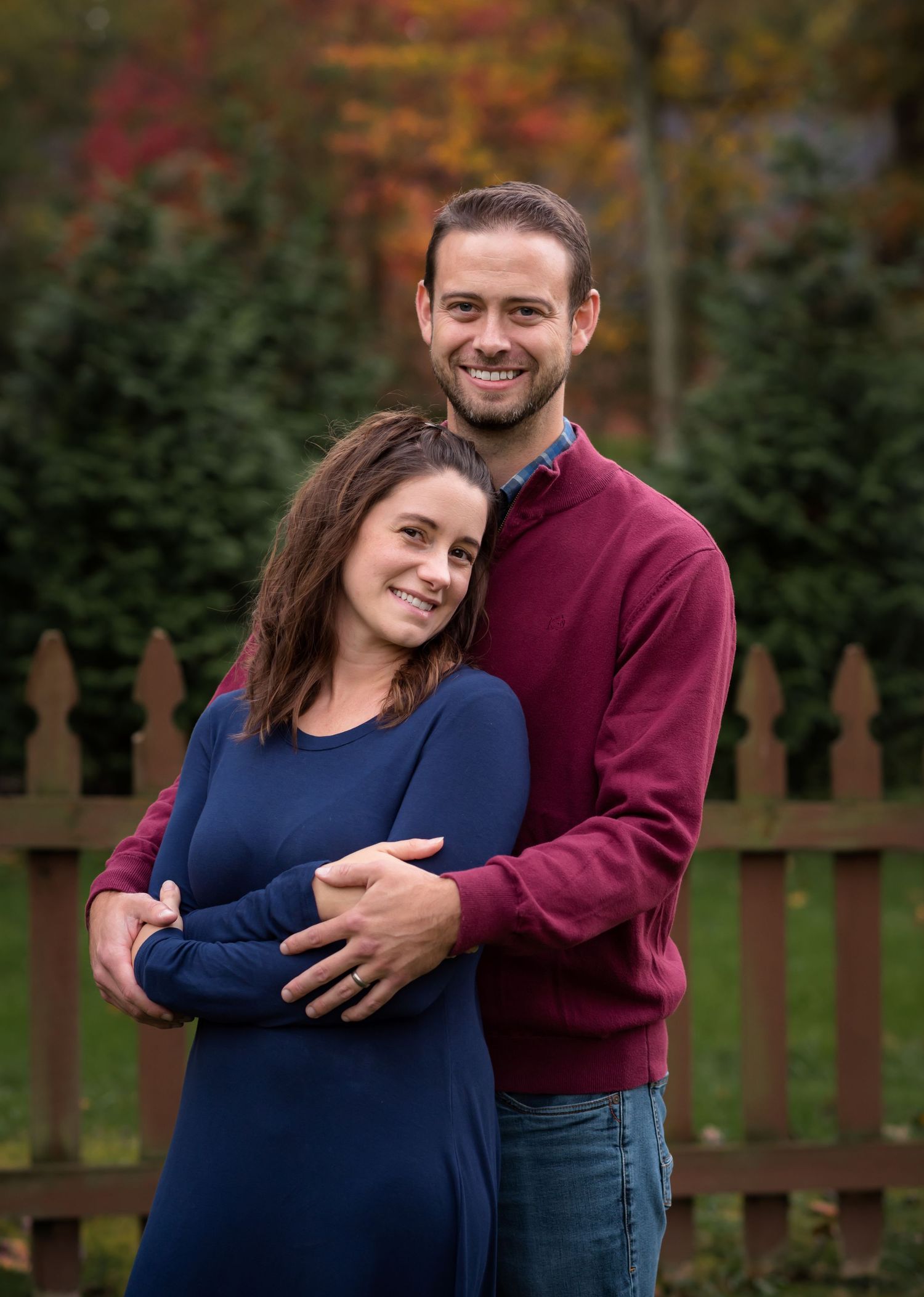 a husband and wife embrace and smile for a portrait with rich fall foliage in the background; shot in Morgantown, WV