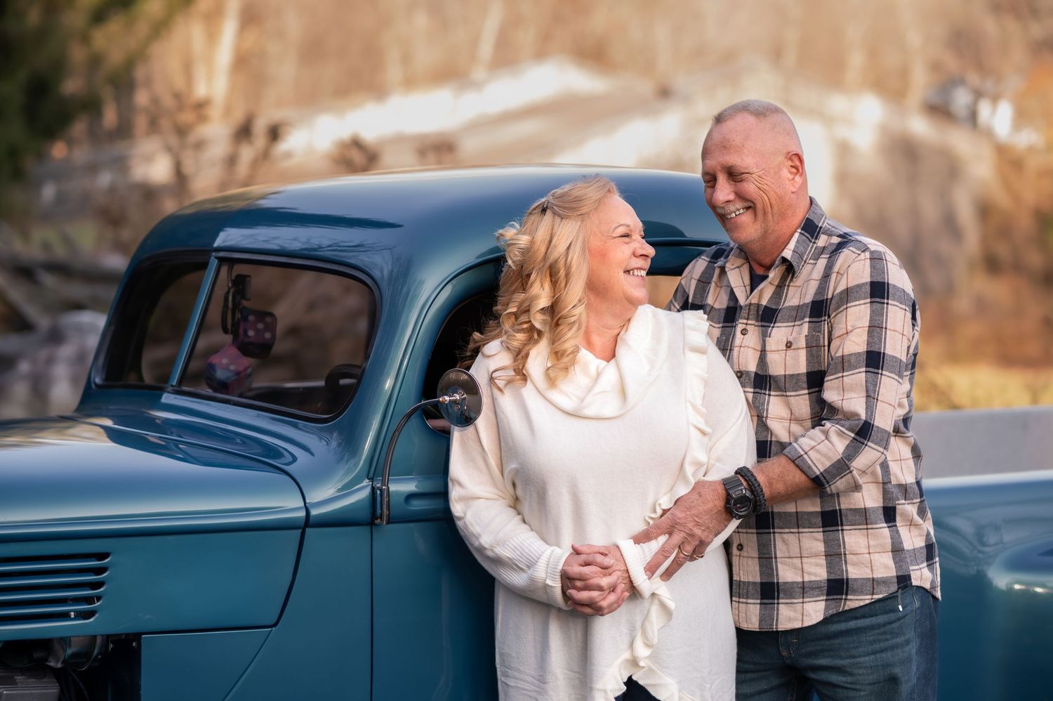 candid portrait of an older couple laughing and embracing in front of a classic model antique truck with fall foliage in the background