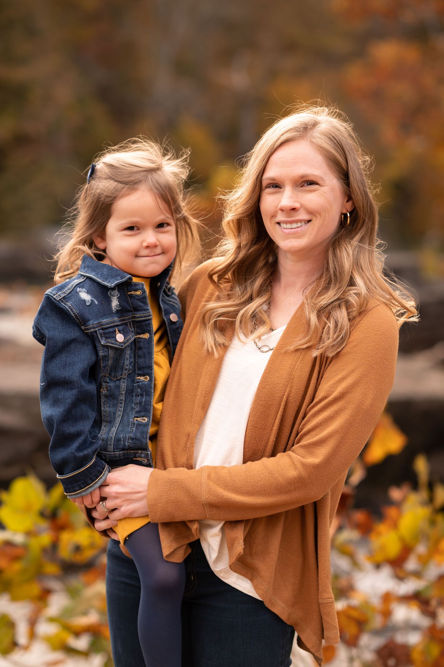 mother holds her young daughter and both smile for a portrait; rocks and waterfall and fall foliage in the background; Valley Falls, WV