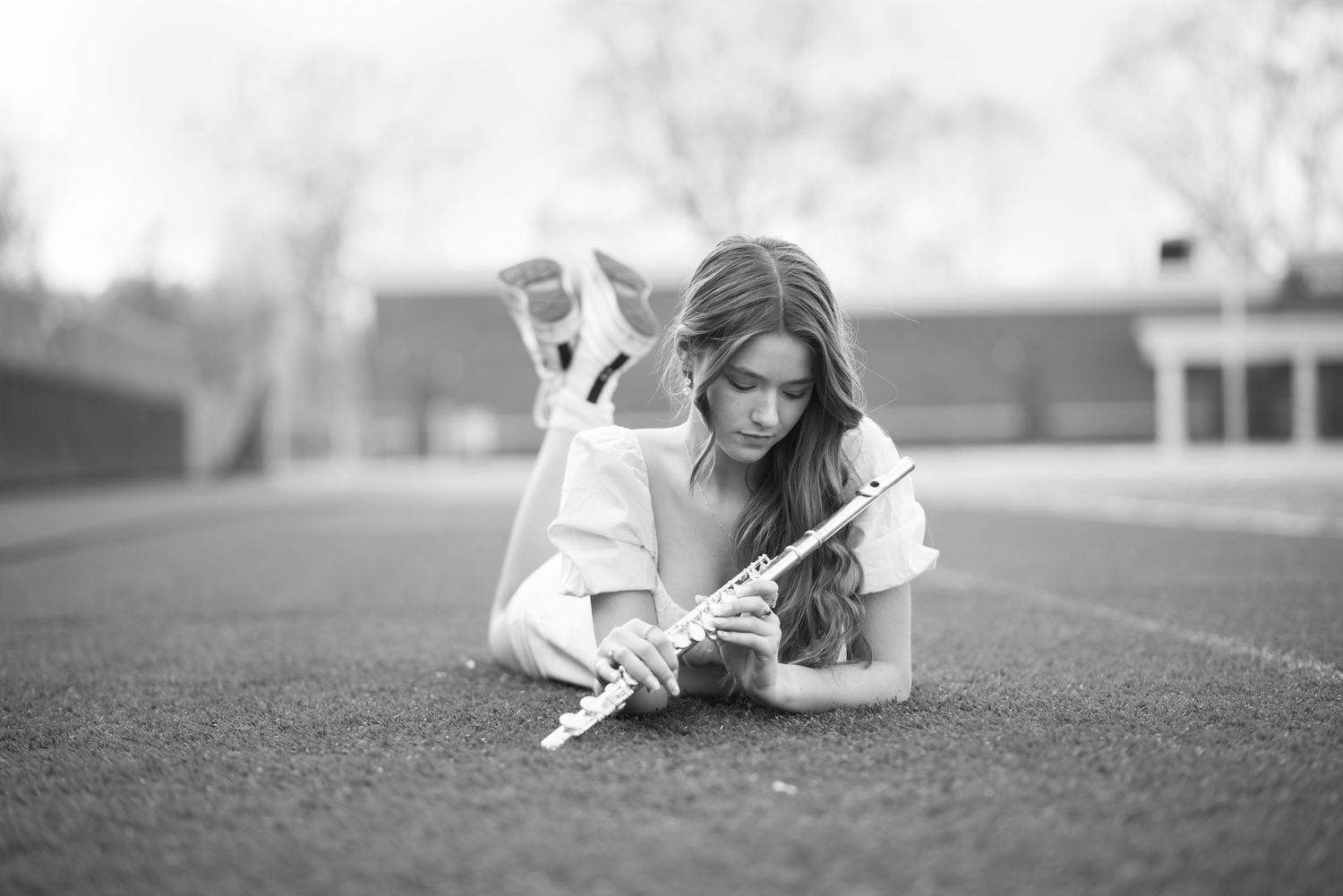 black and white senior portrait of a young woman with brown hair in a white dress, holding her flute and lying on a football field