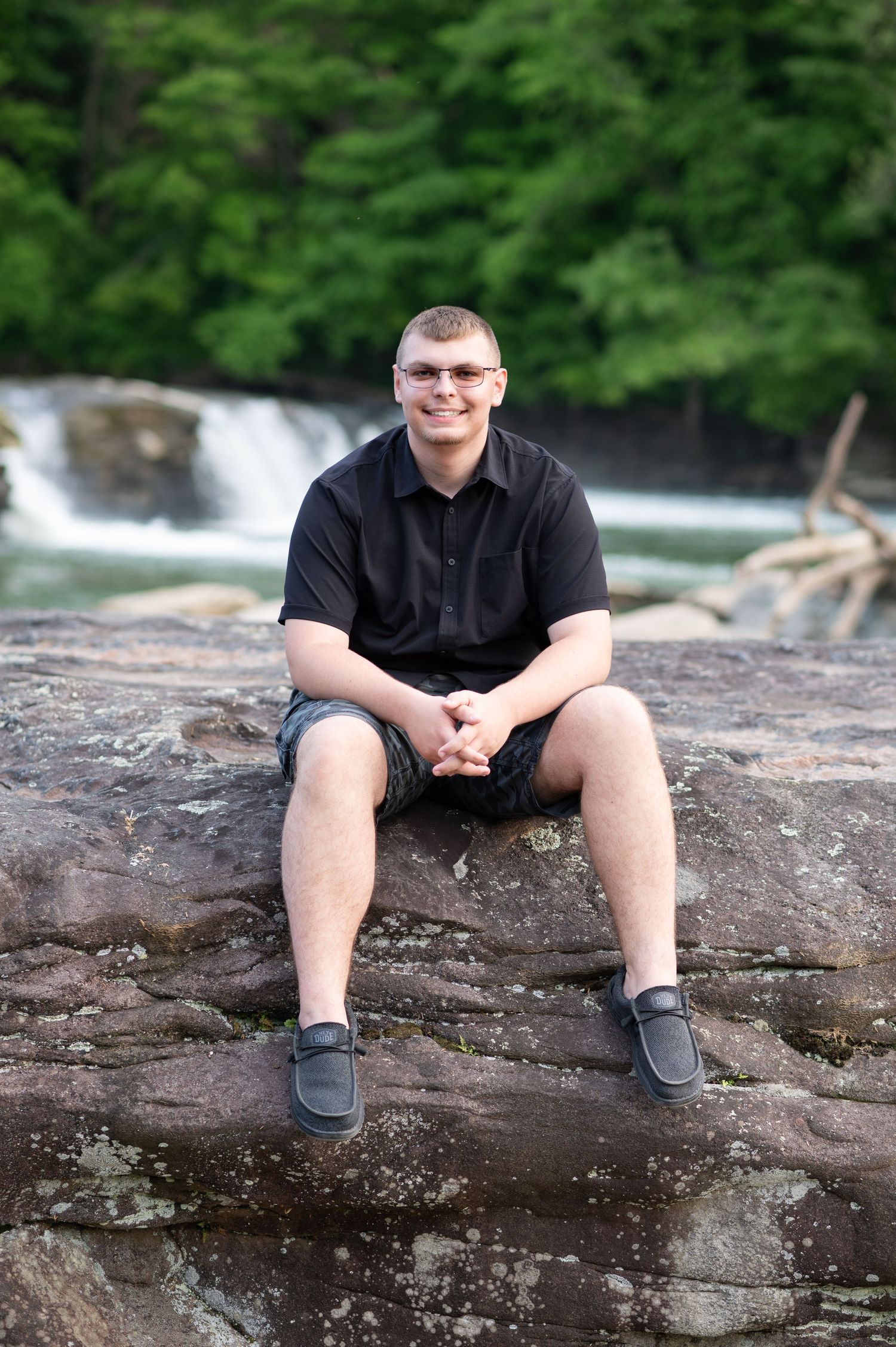 a young high school graduate in black sits on a large rock in front of waterfalls at Valley Falls, WV