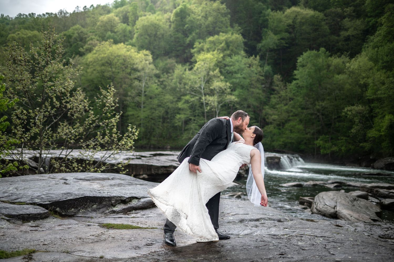 a groom dips his bride and kisses her in the rain in front of waterfalls with woods in the background at Valley Falls State Park, WV