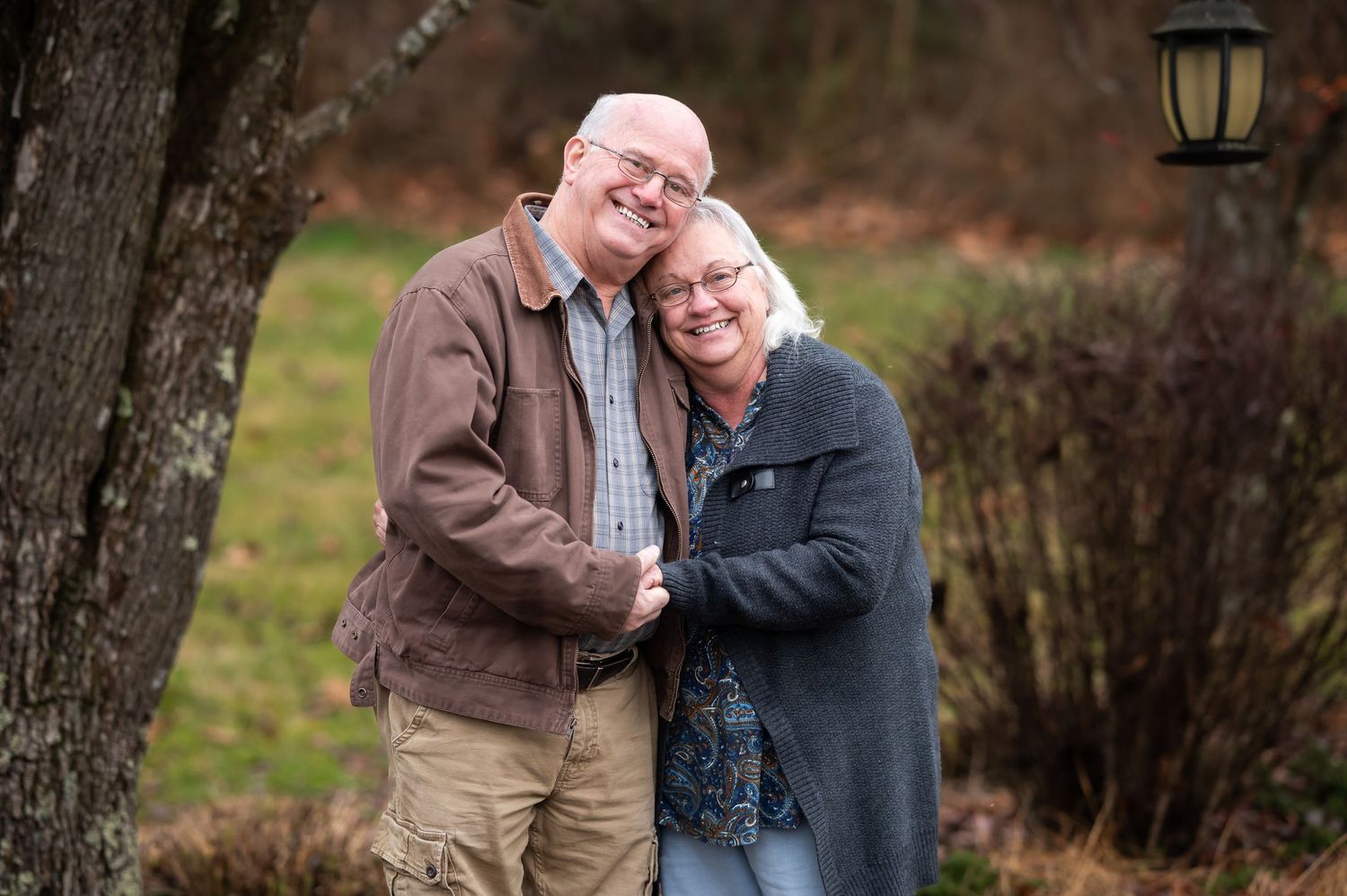 an older couple stand outside under a large tree, holding hands and smiling for a portrait