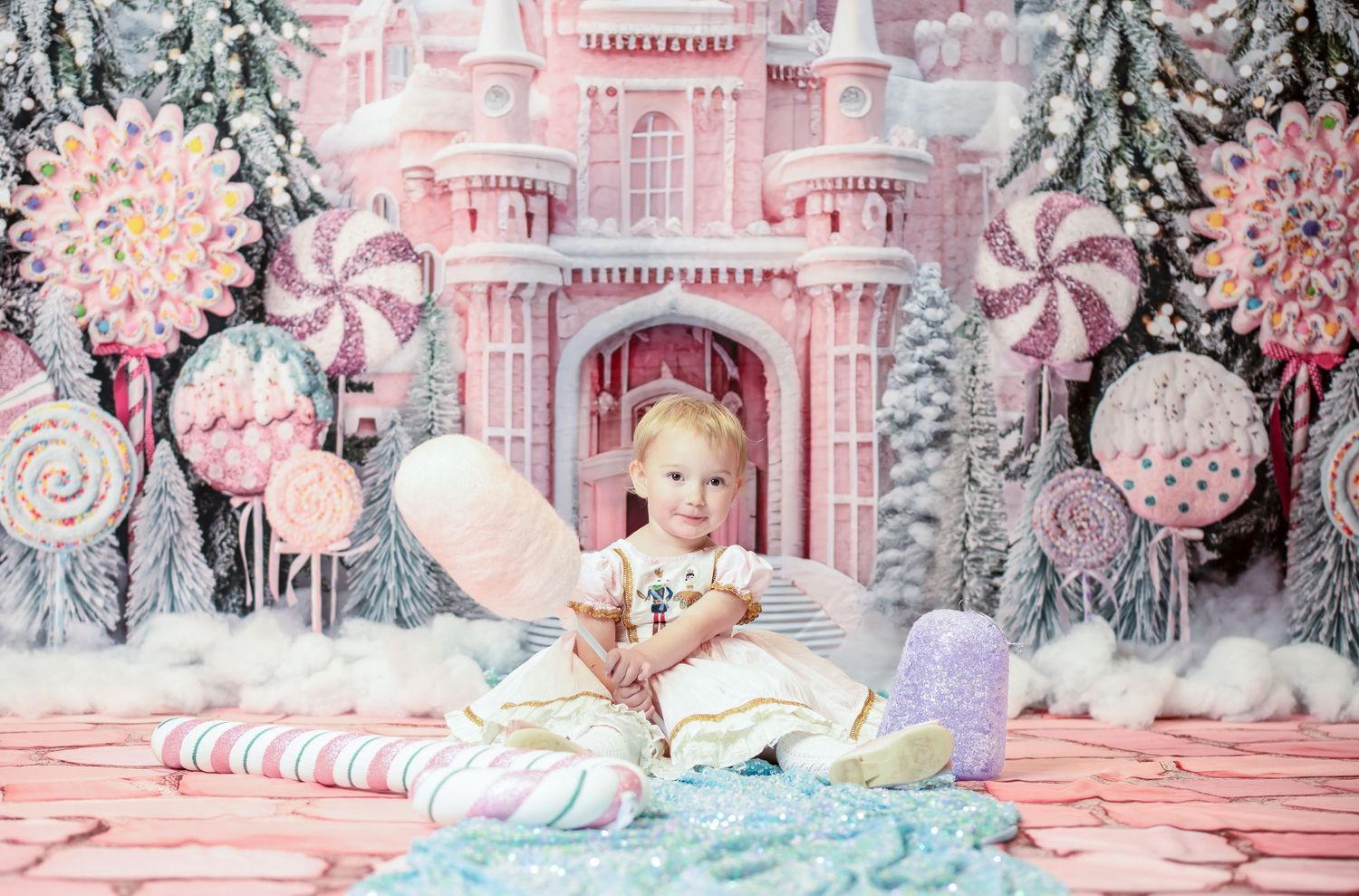 a little girl in a fancy pink and white Christmas dress sits on pink stones with a fanciful pink and white snowy candy castle behind