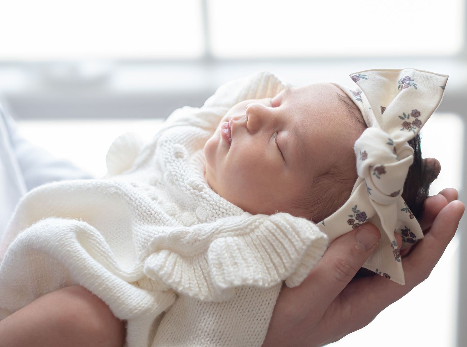 Newborn baby sleeping peacefully in her mother's hands, wearing a white knit outfit and floral bow headband.