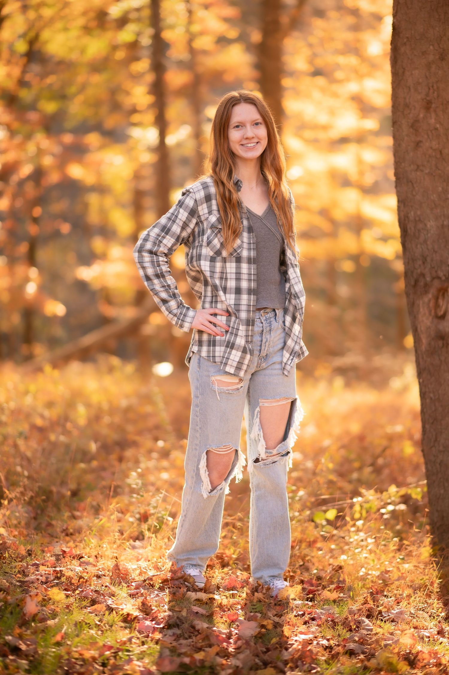 a pretty redheaded senior student in a plaid shirt and ripped jeans poses for a portrait in front of beautiful fall scenery in Morgantown WV