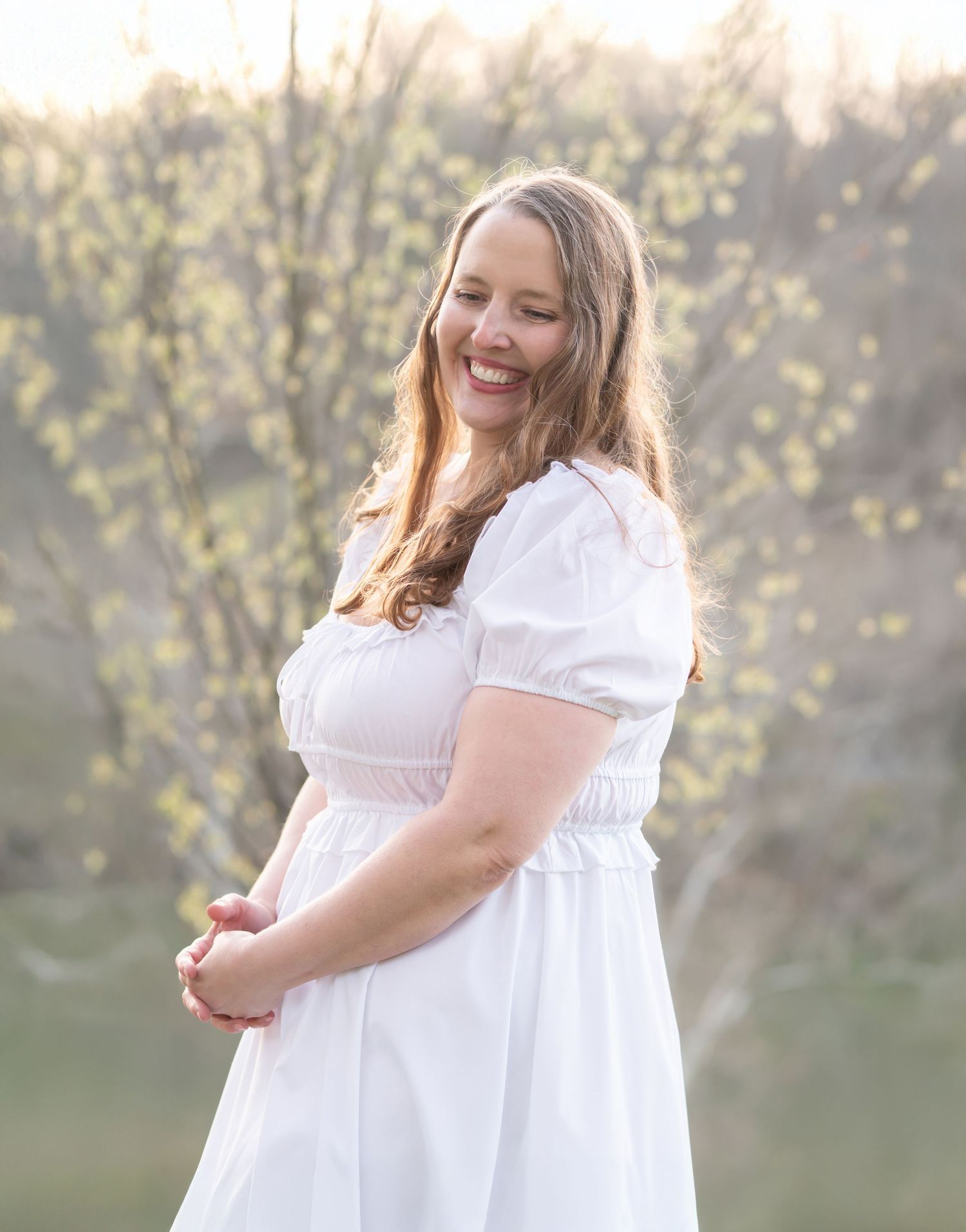 a woman with long brown hair in a white dress poses for a portrait and laughs, with fresh spring greenery and a river behind her