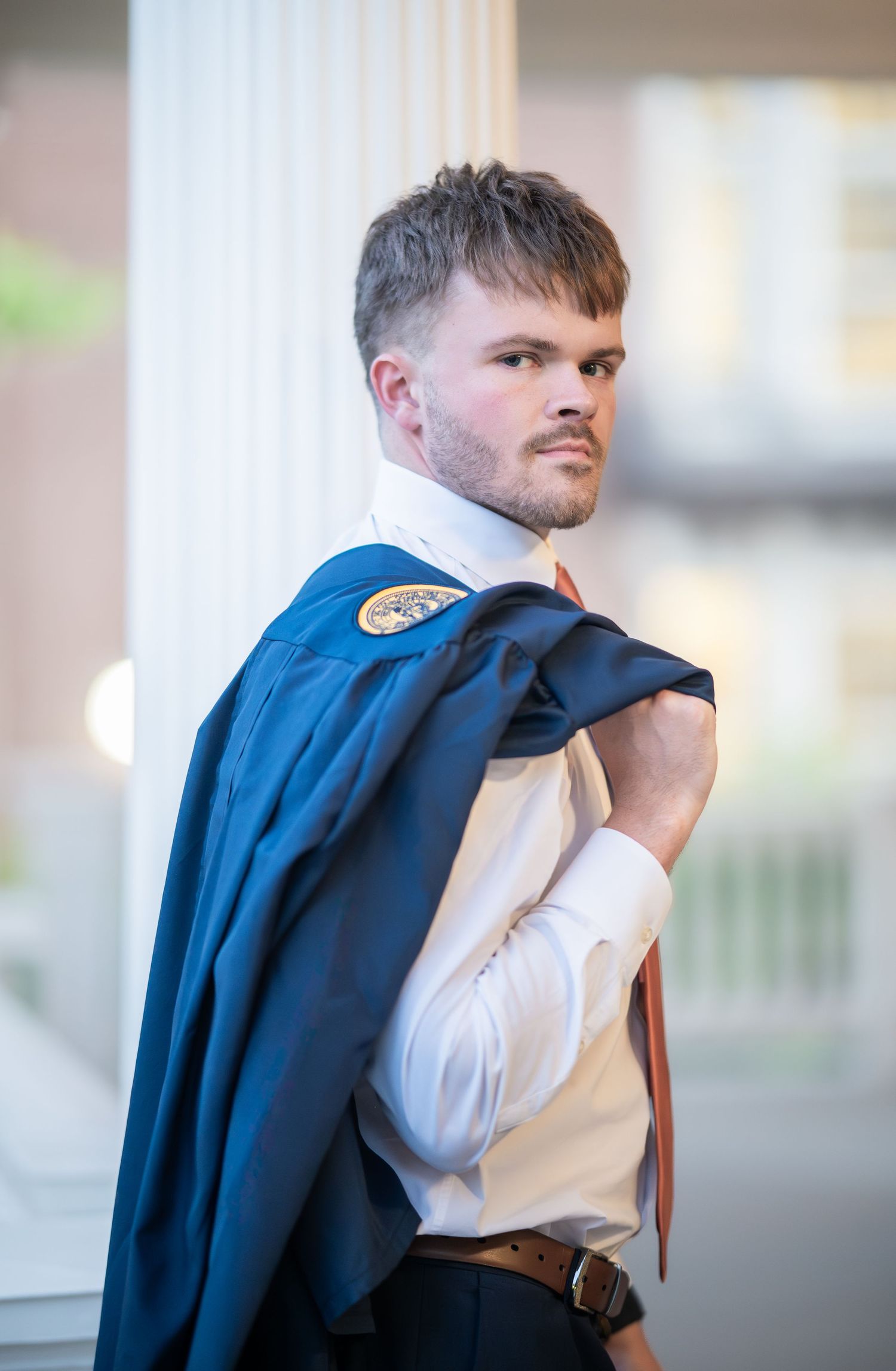A WVU graduate in a blue suit and red tie poses for a portrait with his graduation gown draped over his shoulder, looking back at the camera