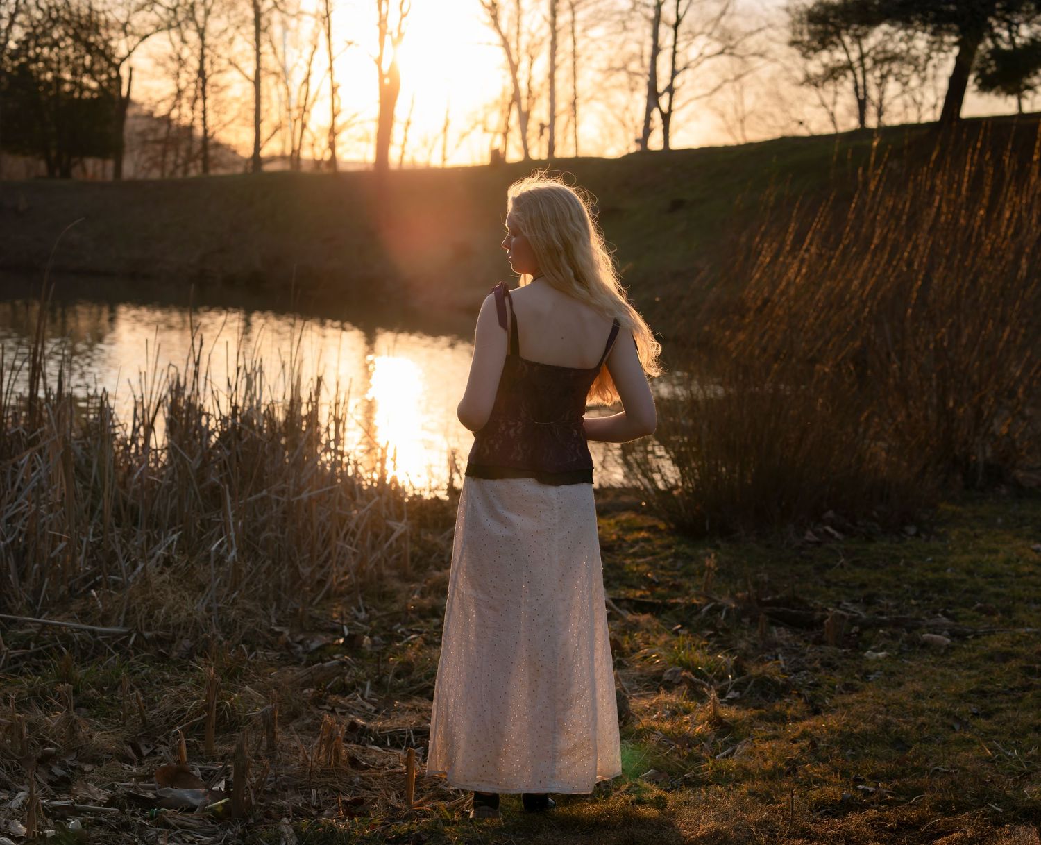 environmental portrait of a young woman with long blonde hair, standing and looking over a sunlit lake at sunset
