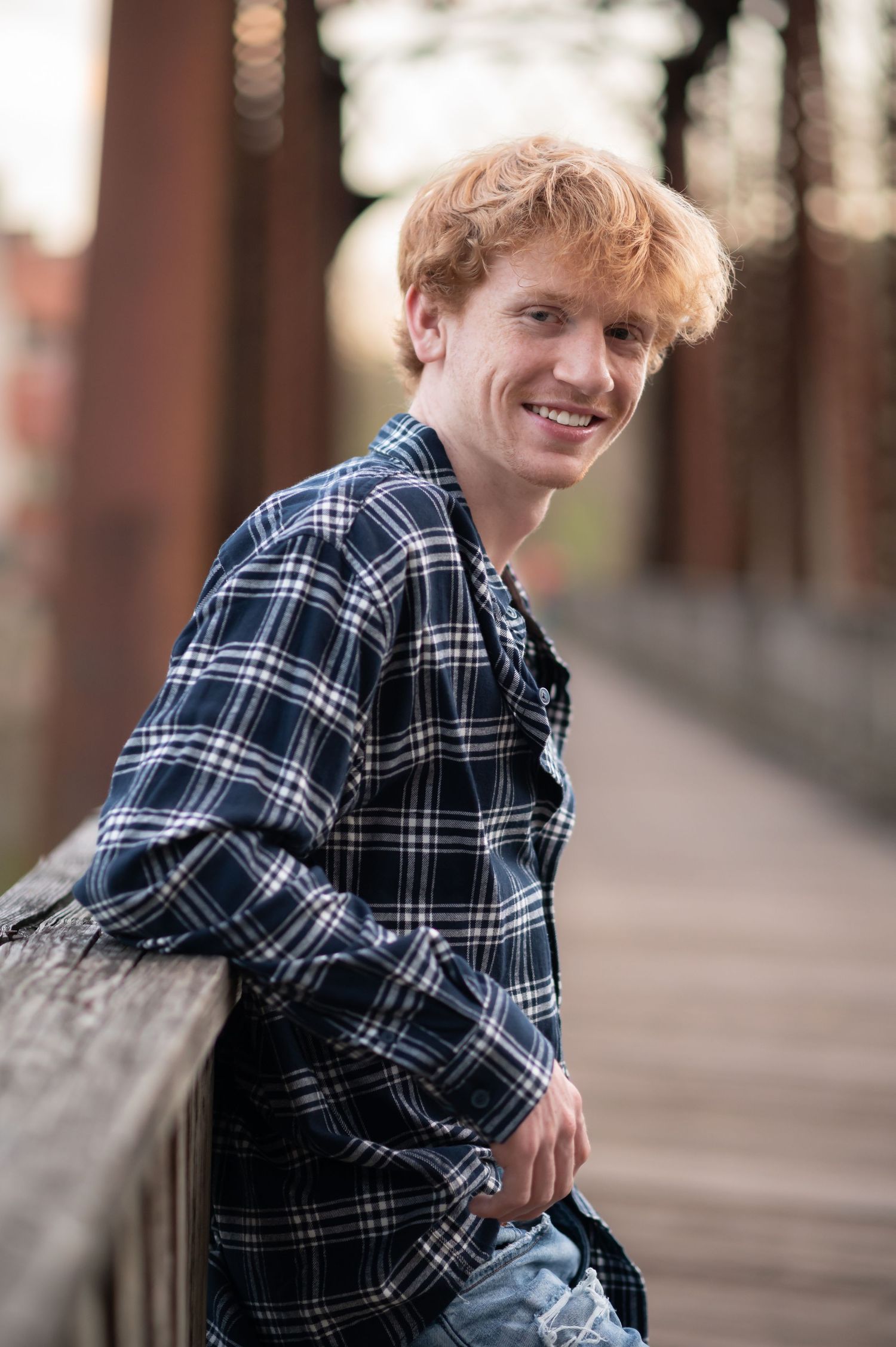 a curly-haired student in a plaid shirt and jeans lounges against the railing of an old wooden and iron bridge in downtown Morgantown, WV