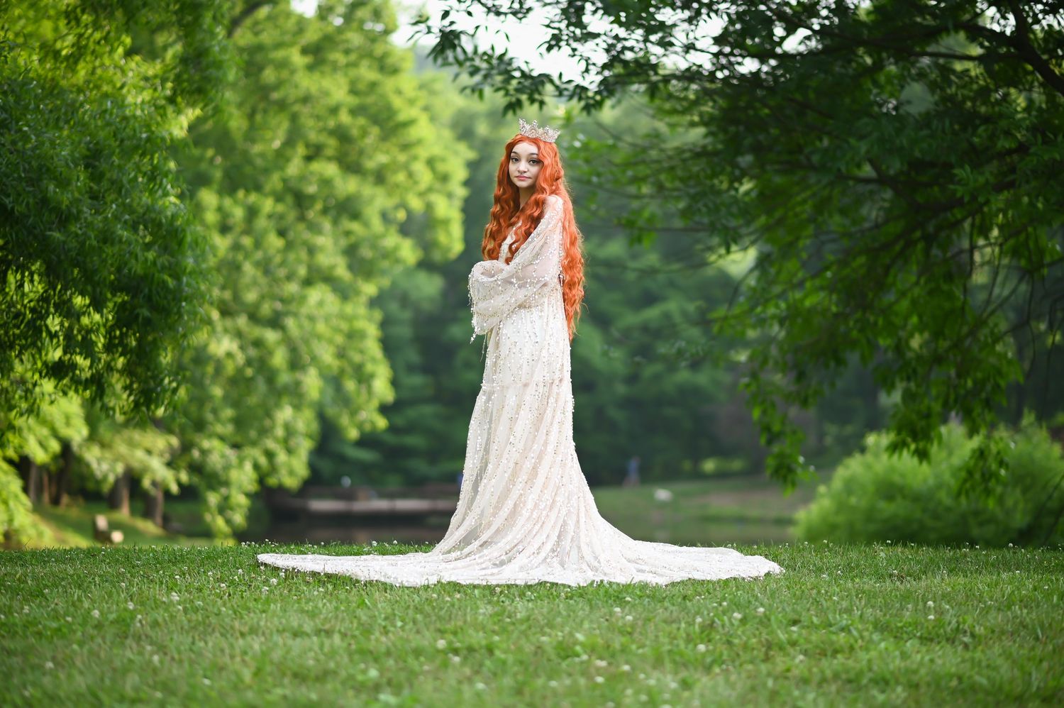 a young woman with very long red hair in a white gown and crown poses in the green grass in front of rich green trees in Morgantown, WV