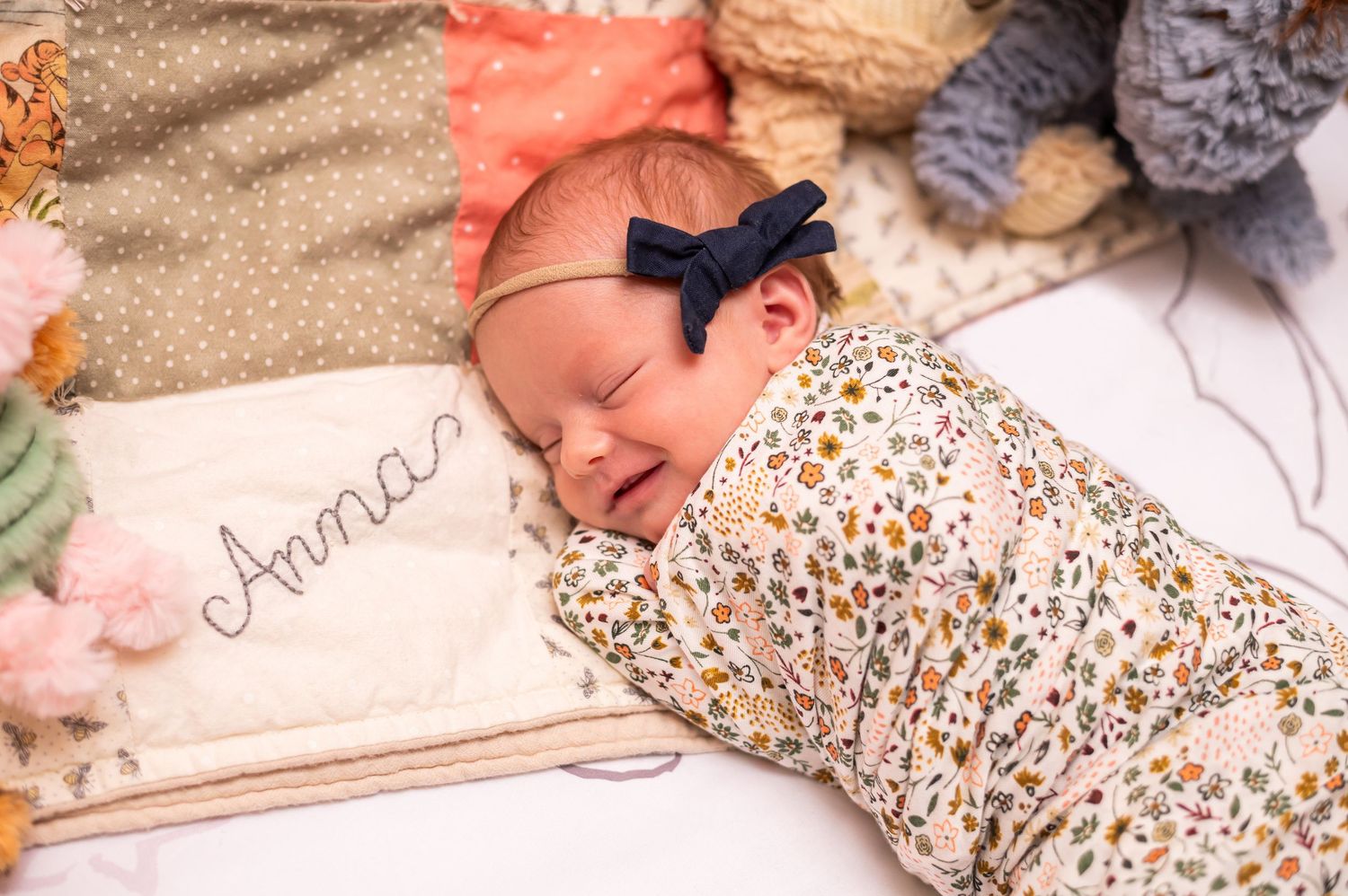 Newborn baby girl named Anna swaddled in floral wrap, wearing navy bow headband, sleeping peacefully on personalized blanket.