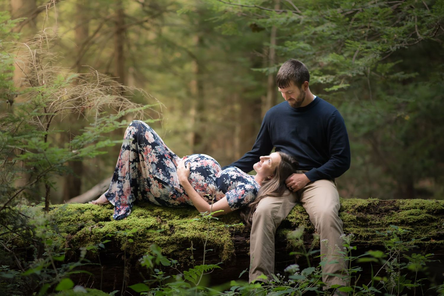 a pregnant woman in a floral dress poses on an old fallen log with her husband in the middle of the woods, with sunlight falling around them