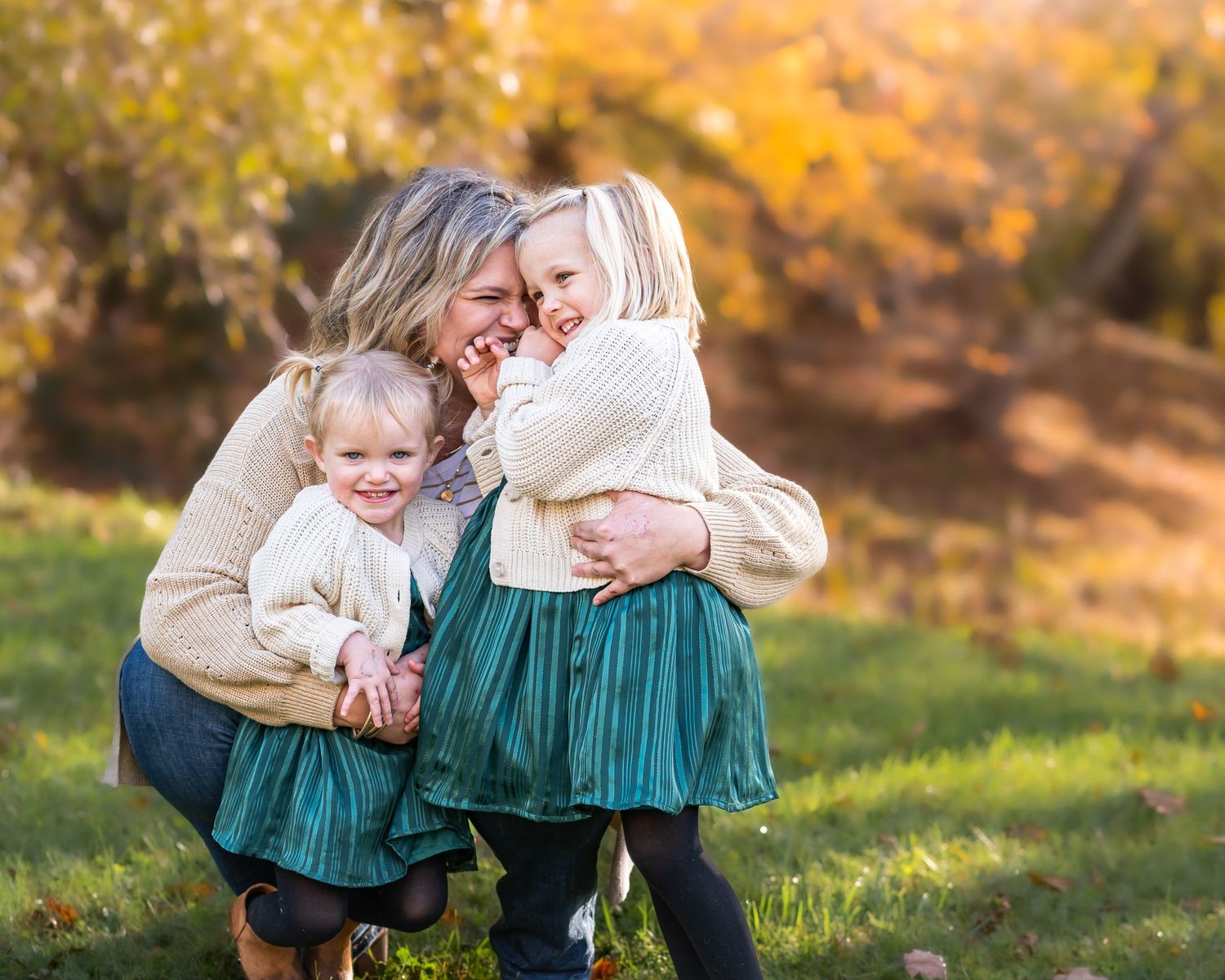 a mother stoops to hug her two little blonde daughters as they all laugh, with autumn scenery in the background