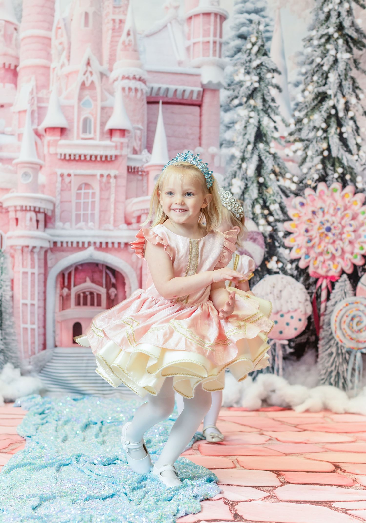 a little girl in a shimmering pink and white Christmas dress smiles and twirls with an elaborate pink candy castle in the background