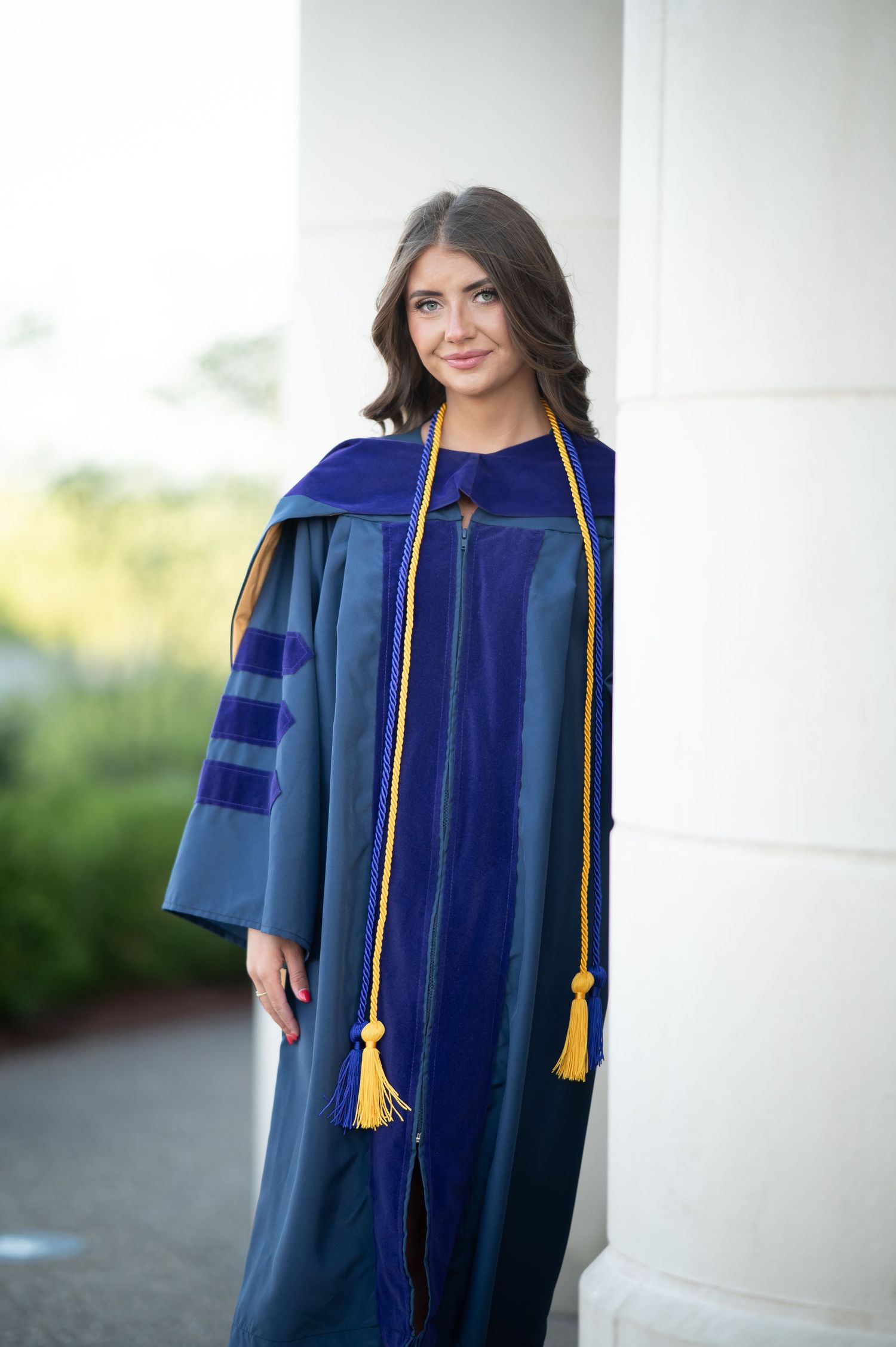 a WVU graduating student leans against a large white pillar while wearing her cap and gown