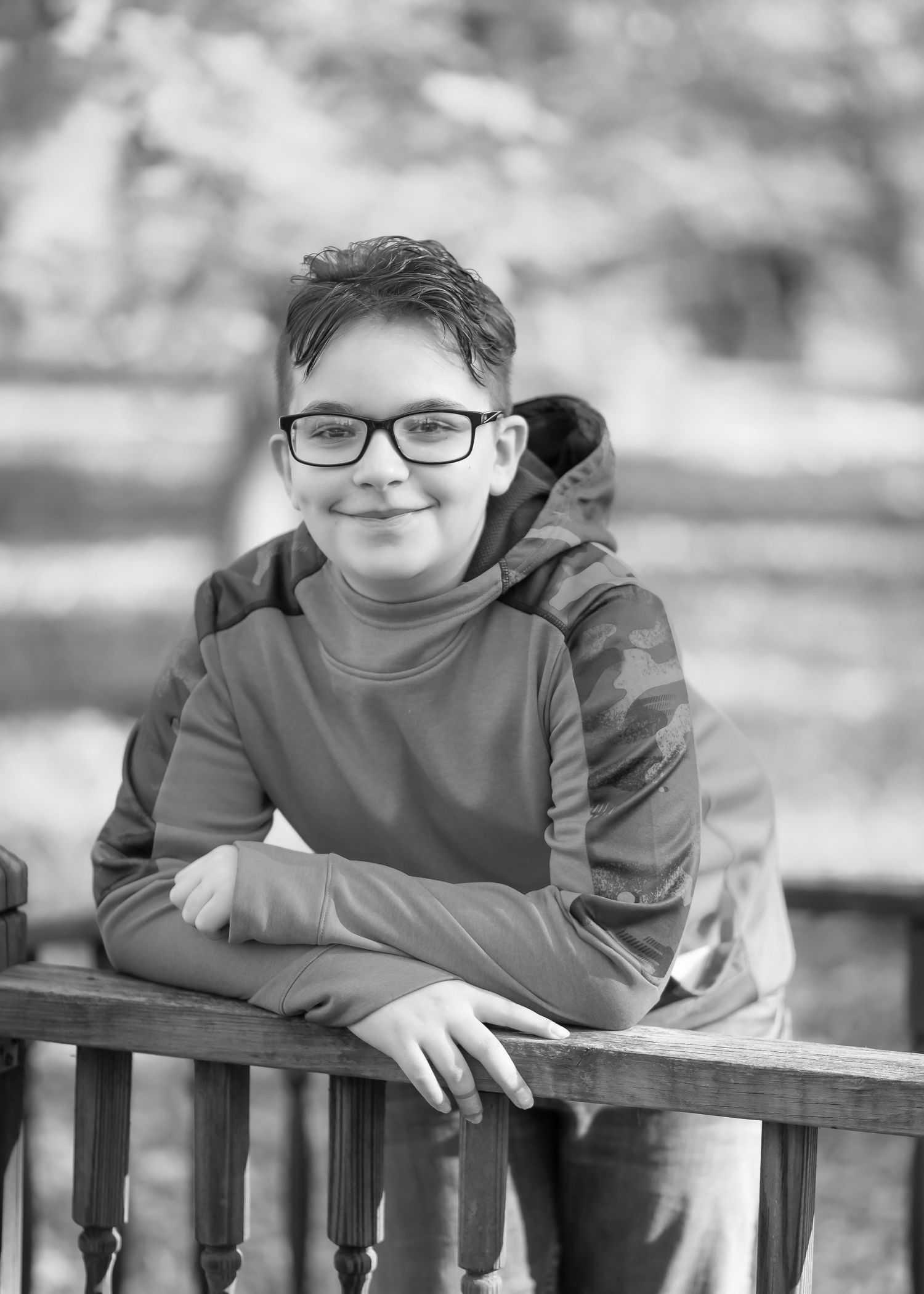 a young boy with dark hair and glasses leans on an old wood railing and smiles into the camera for a portrait
