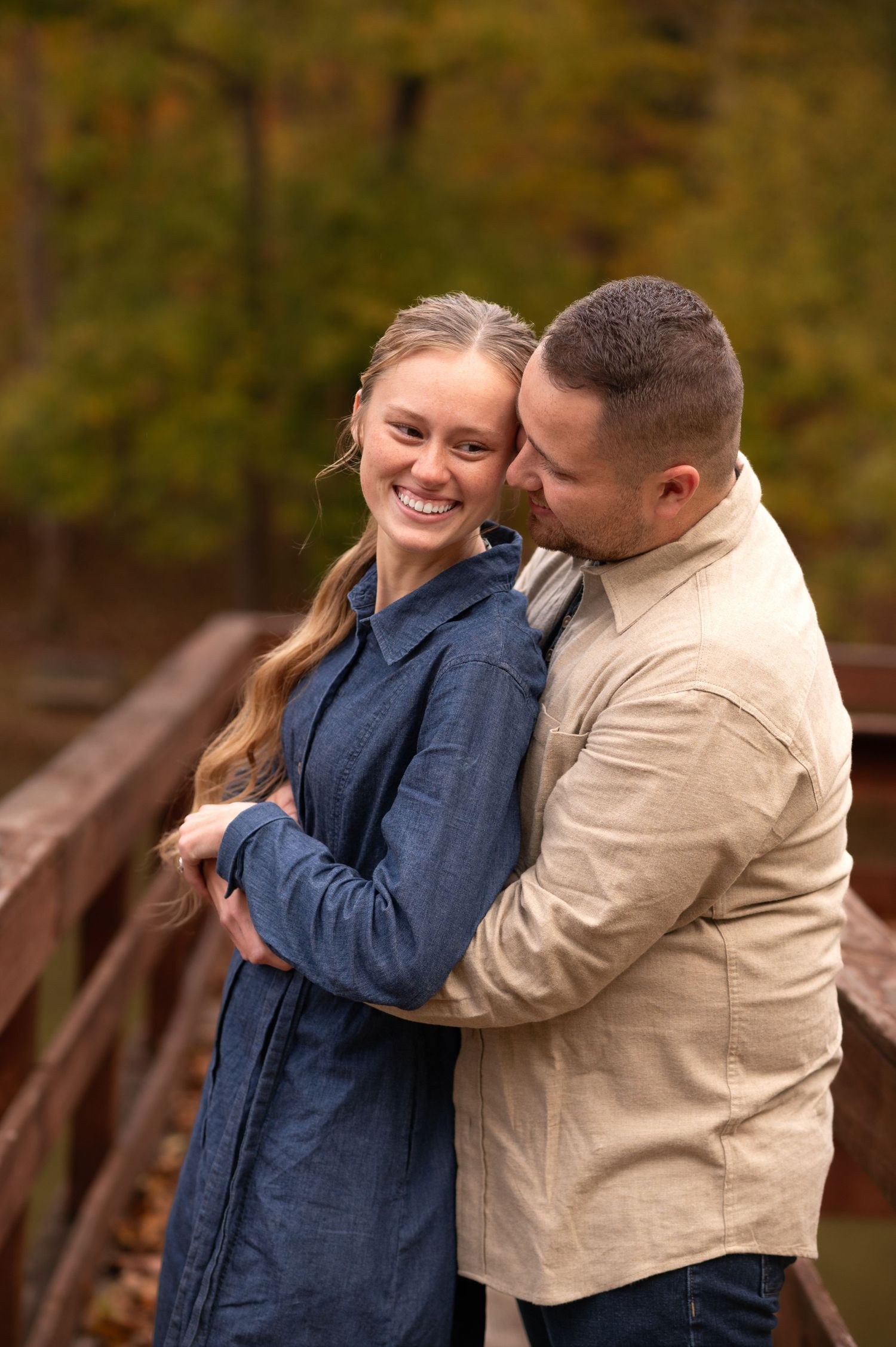 a sweet young couple in autumn outfits leans against a wooden railing, embracing and smiling