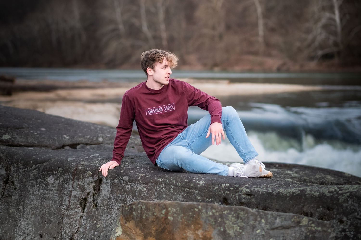 a high school graduate in a maroon top and jeans strikes a stylish seated pose on a large rock with waterfalls, a river and trees behind