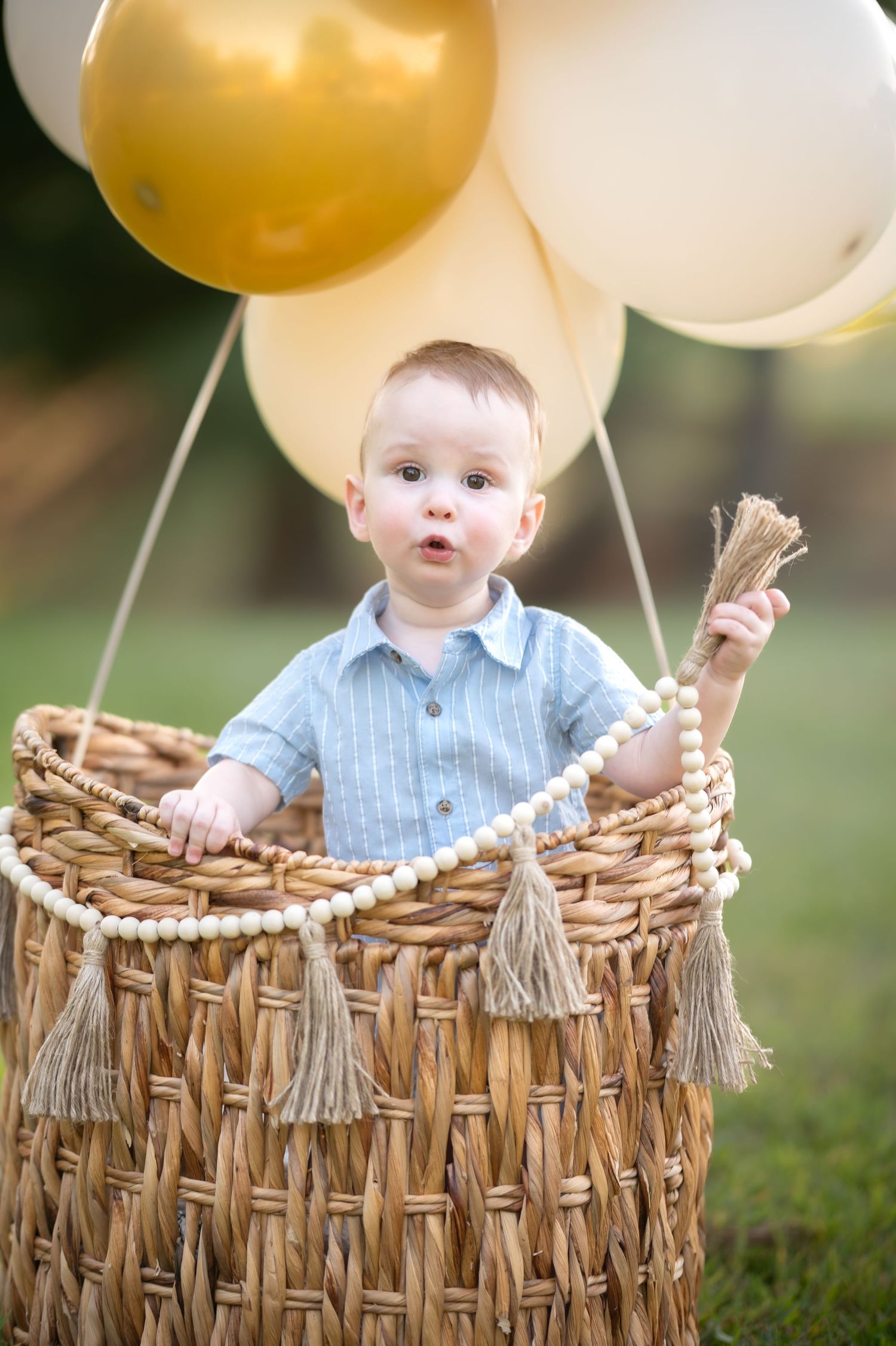 a little boy in a blue romper stands in a little hot air balloon basket with many gold and white balloons over his head