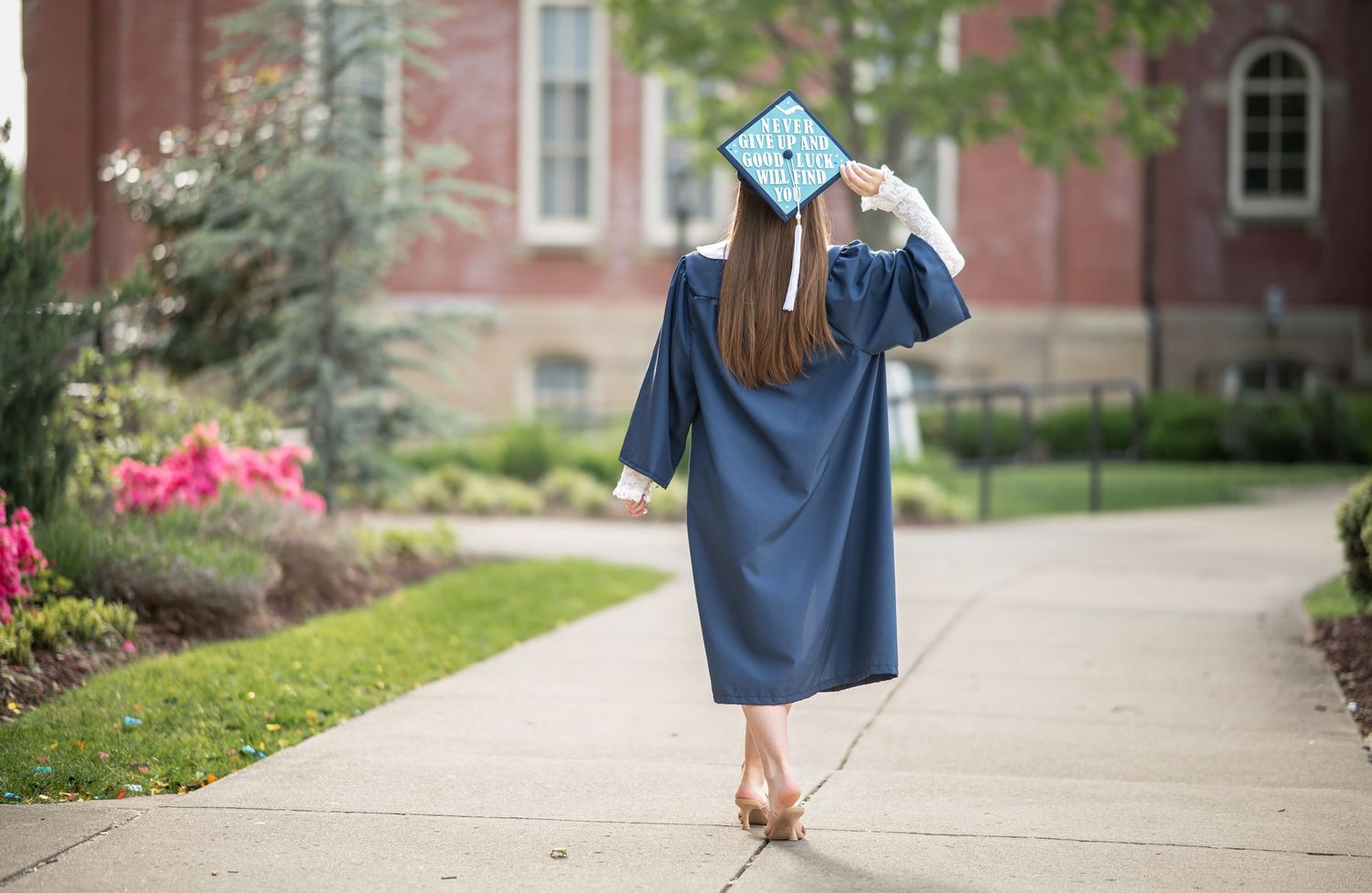 a WVU grad student in cap and gown walks away across the lawn in Woodburn Circle with Woodburn Hall in the background, in Morgantown, WV