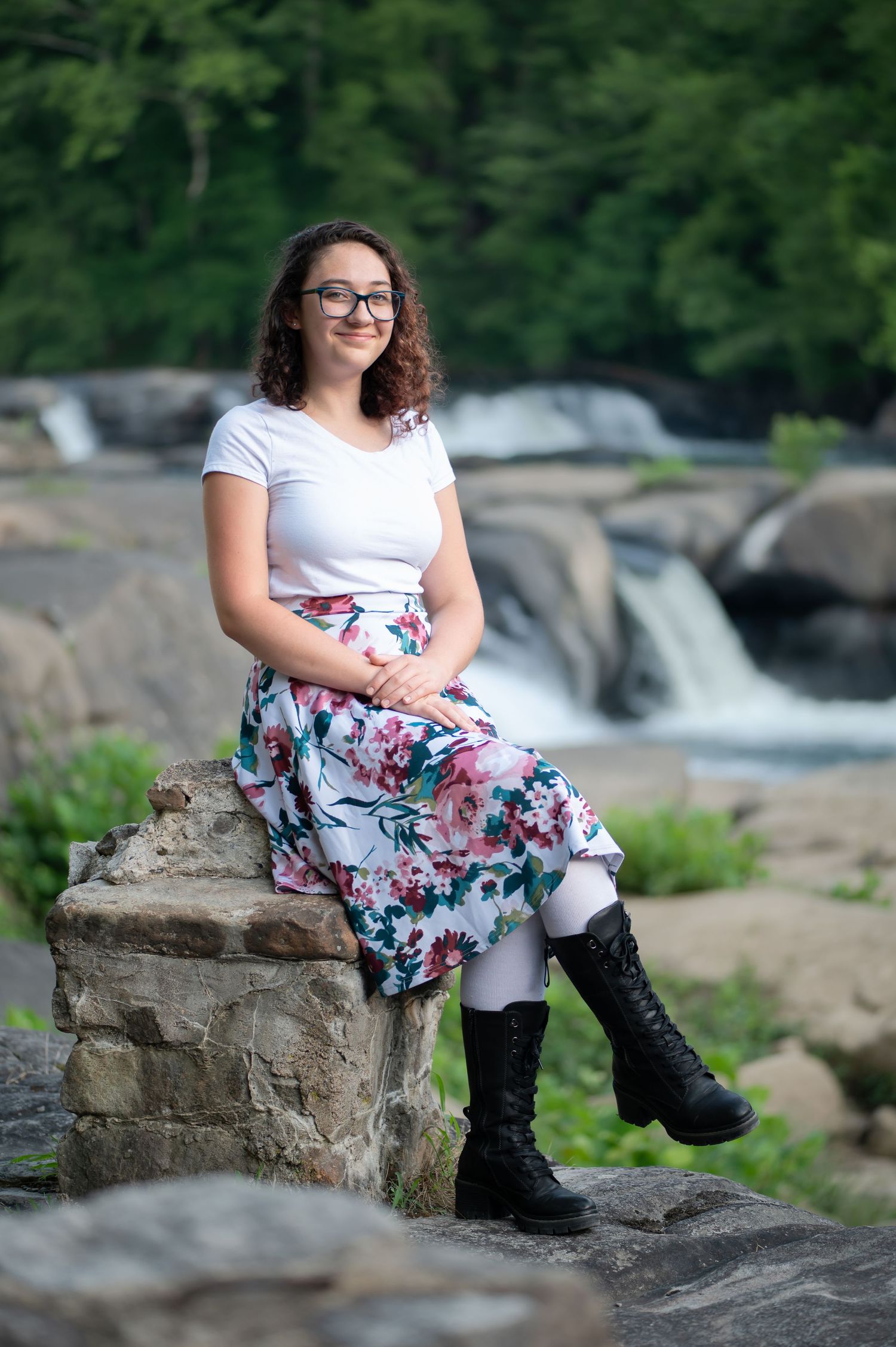a dark haired young woman in a white top and floral skirt poses for a portrait on a large rock in front of waterfalls