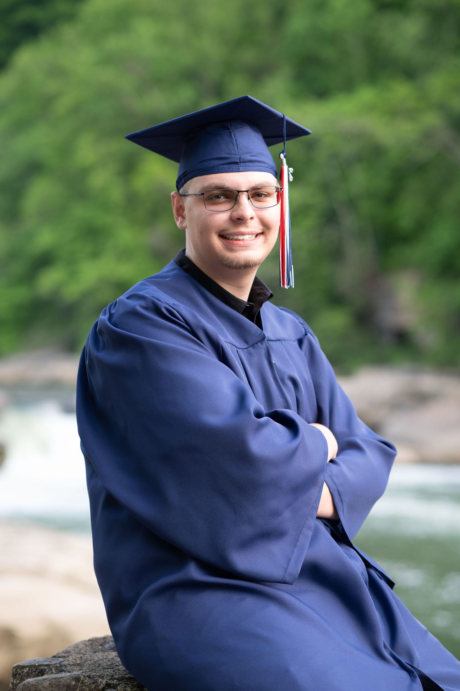 a young man in a navy blue cap and gown poses for a senior portrait in front of a river at Valley Falls State Park in Morgantown, WV